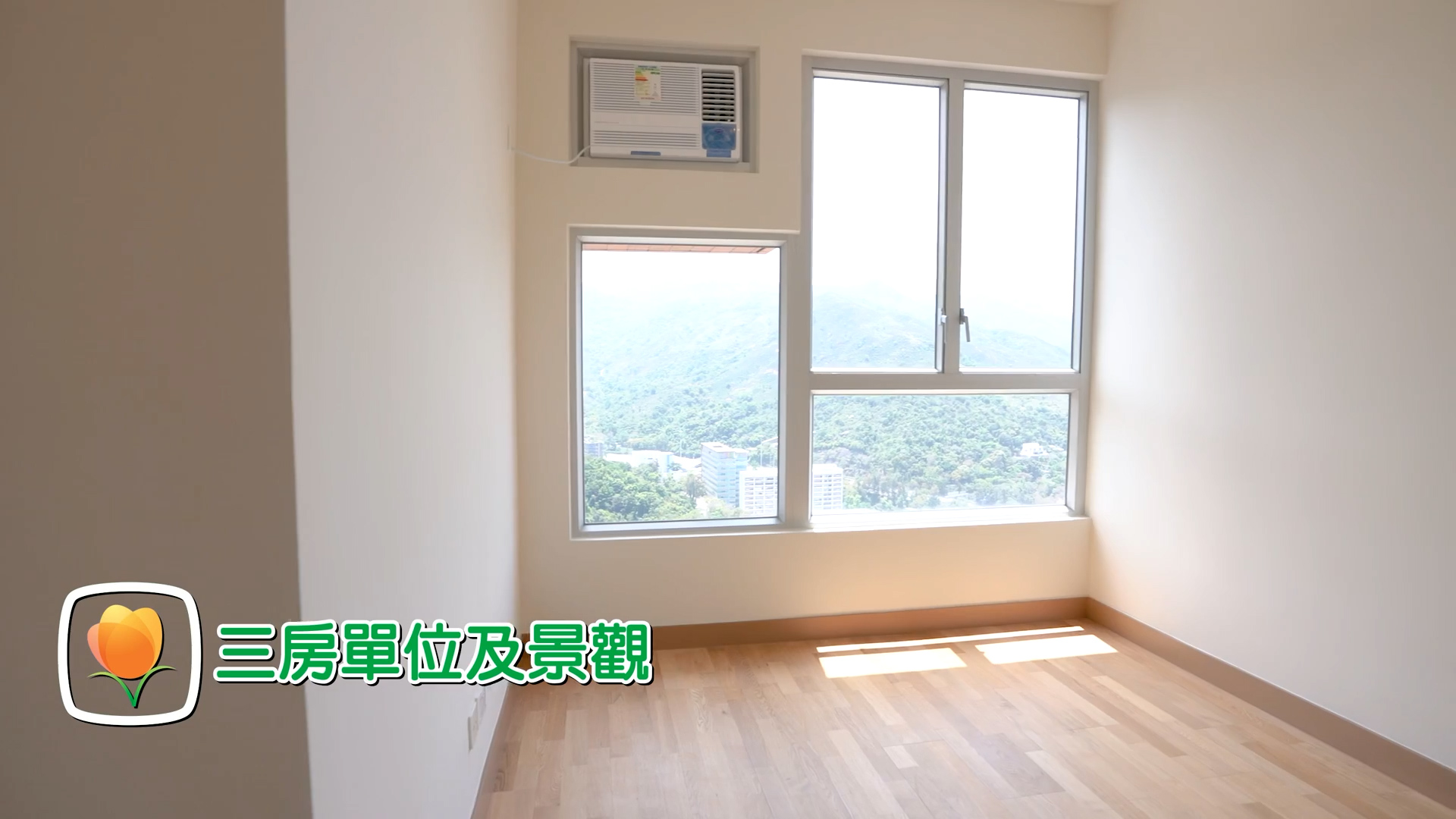 A bright, empty room with light wood flooring, large windows showing a scenic view of mountains and buildings, an air conditioner above the window, and Chinese text with a flower graphic in the lower left corner.