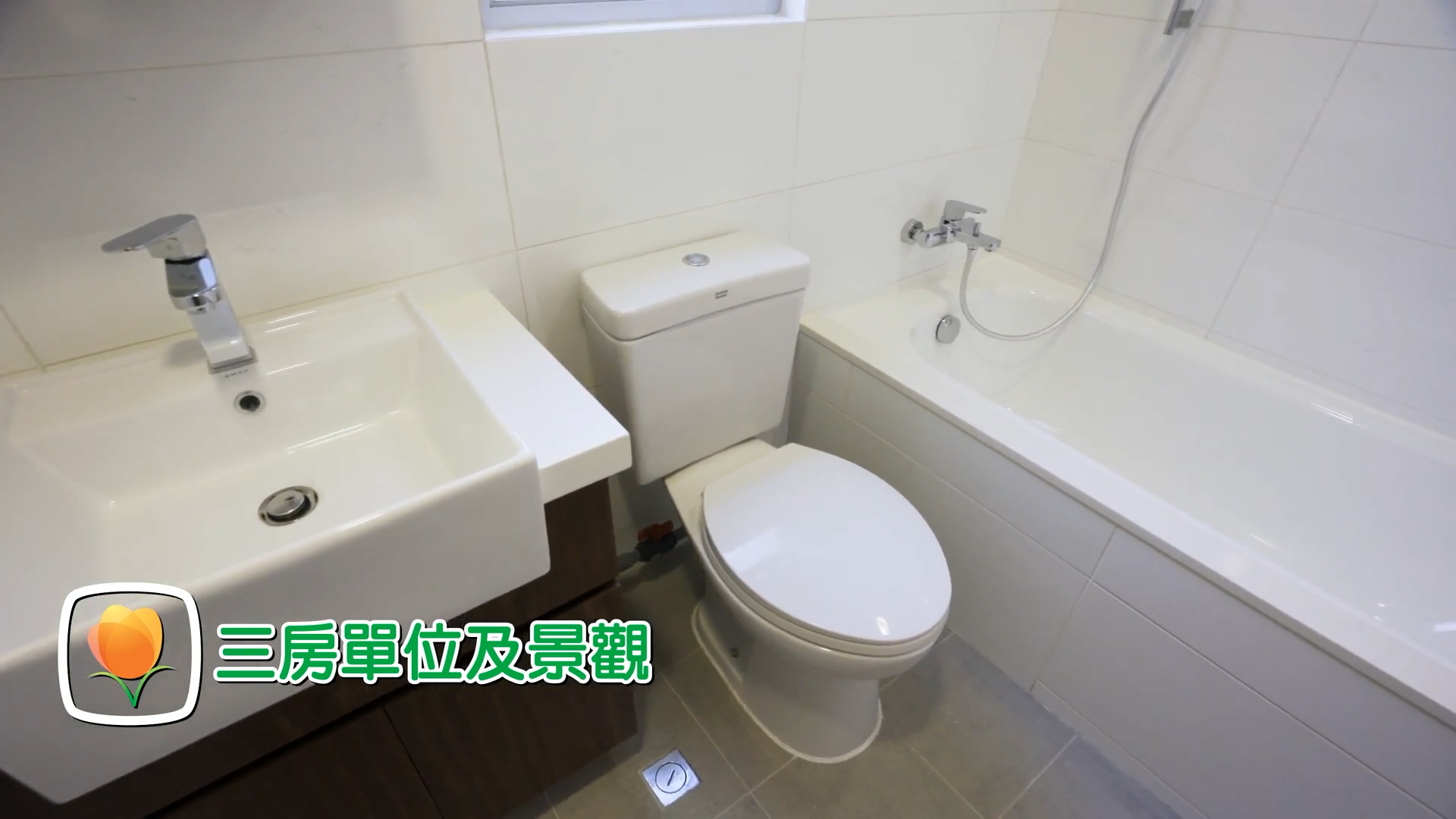 A modern bathroom with a white sink, toilet, and bathtub. The walls and floor are tiled. There is a faucet above the sink and a handheld shower above the tub. Text in Chinese and an orange flower icon appear on the image.