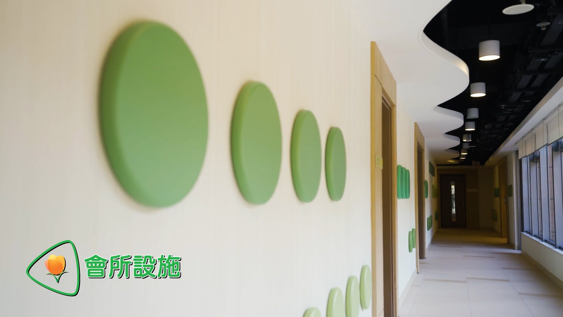 A hallway with green circular decorations on a cream-colored wall, doors along the right side, and windows letting in natural light. Text in Chinese and a logo appear in the lower left corner.
