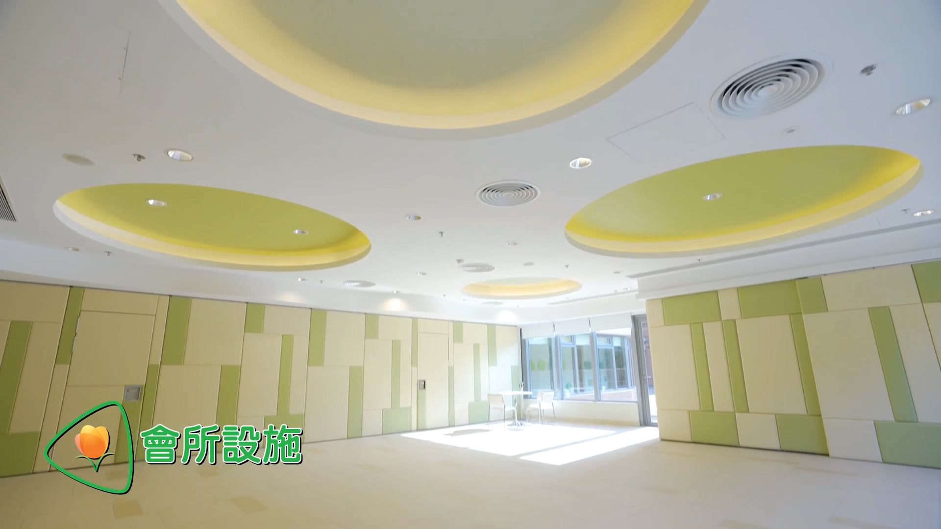 A spacious, modern room with circular ceiling lights, geometric wall panels in soft green and beige tones, and a small table with chairs near large windows. Green Chinese text and a peach icon appear in the lower left corner.