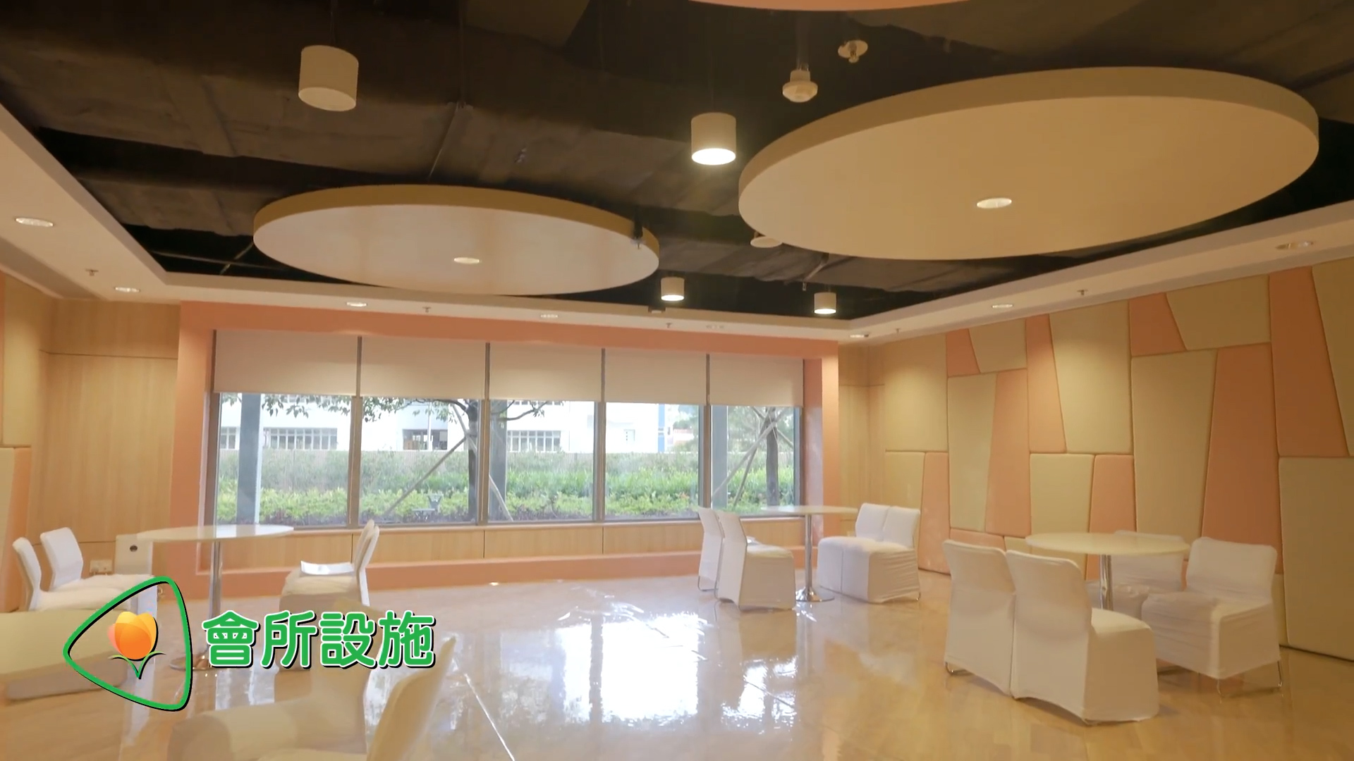 A modern lounge with round ceiling panels, cream and peach walls, large windows showing greenery outside, and several white-covered chairs and tables. Chinese text and an icon appear in the lower left corner.
