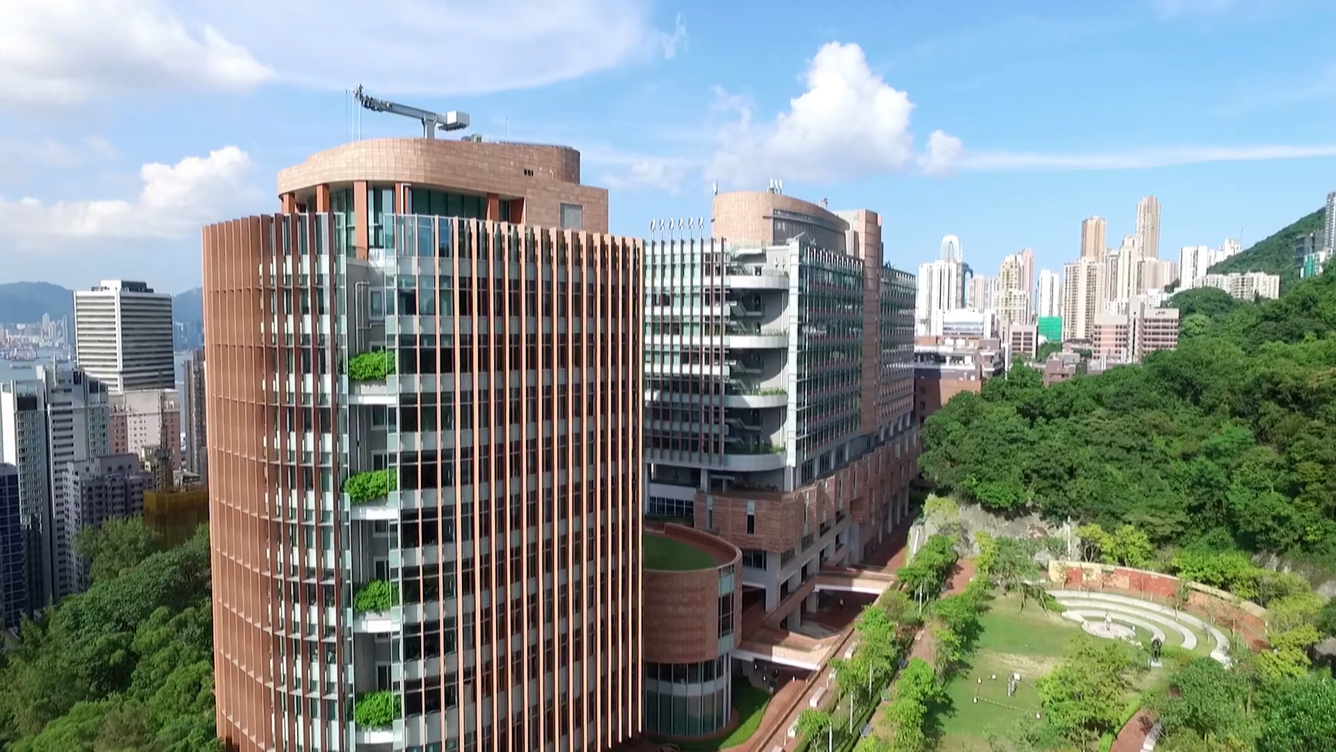Aerial view of modern, multi-story brick and glass buildings surrounded by greenery, with a city skyline in the background and a clear blue sky above.