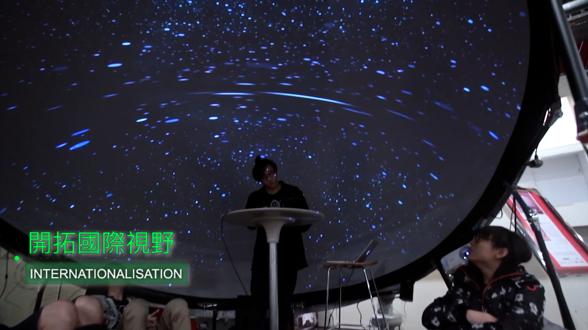 A person stands at a podium in front of a large dome screen displaying a swirling starfield, while others sit nearby. Text in Japanese and English reads "INTERNATIONALISATION.
