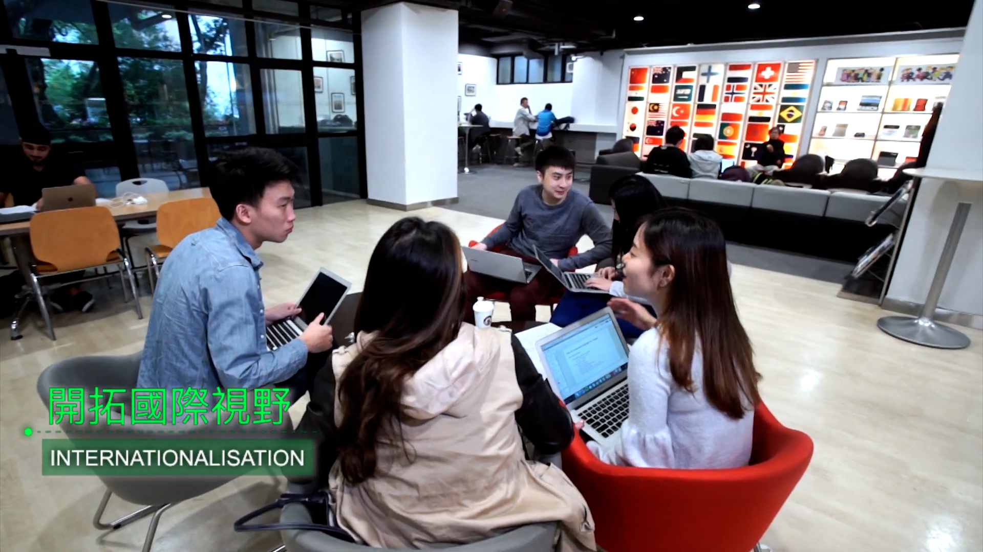 Four students sit in a circle with laptops, talking and collaborating in a modern lounge area. In the background, people work and international flags are displayed on the wall. Text reads “INTERNATIONALISATION” in English and Chinese.