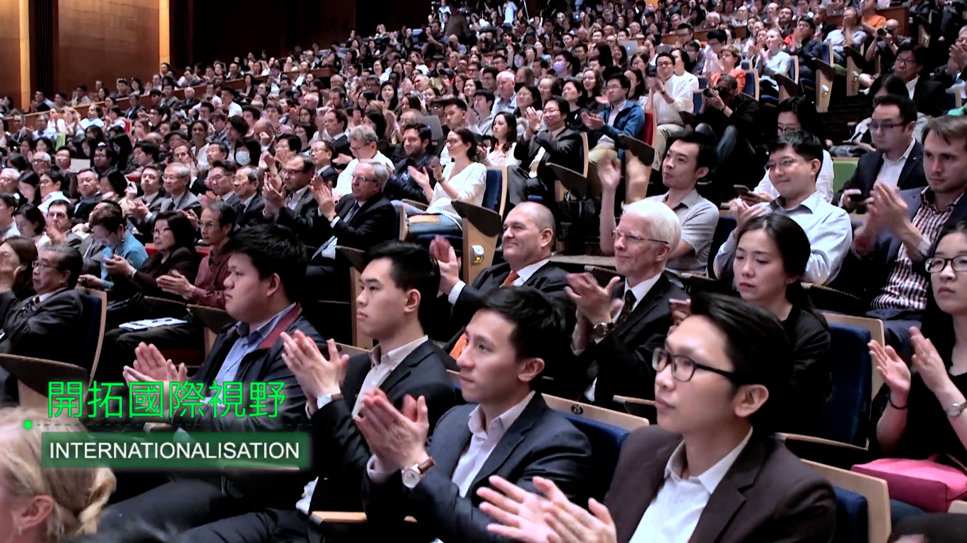 A large audience in business attire sits in an auditorium, attentively watching and applauding. Green and white text on the image reads "INTERNATIONALISATION" and includes Japanese or Chinese characters.
