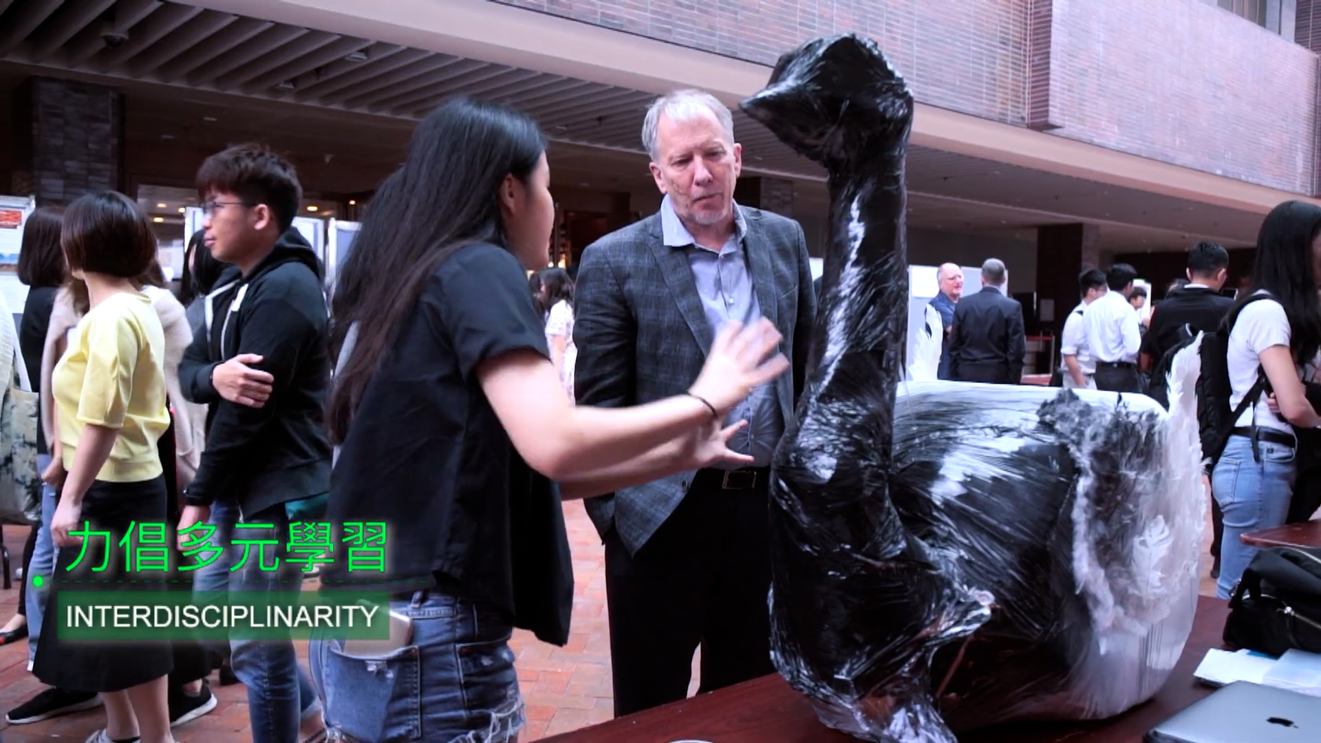 A woman explains a large black sculpture wrapped in plastic to a man at an indoor event, while other people interact in the background. Overlay text reads “INTERDISCIPLINARITY” in English and Chinese.