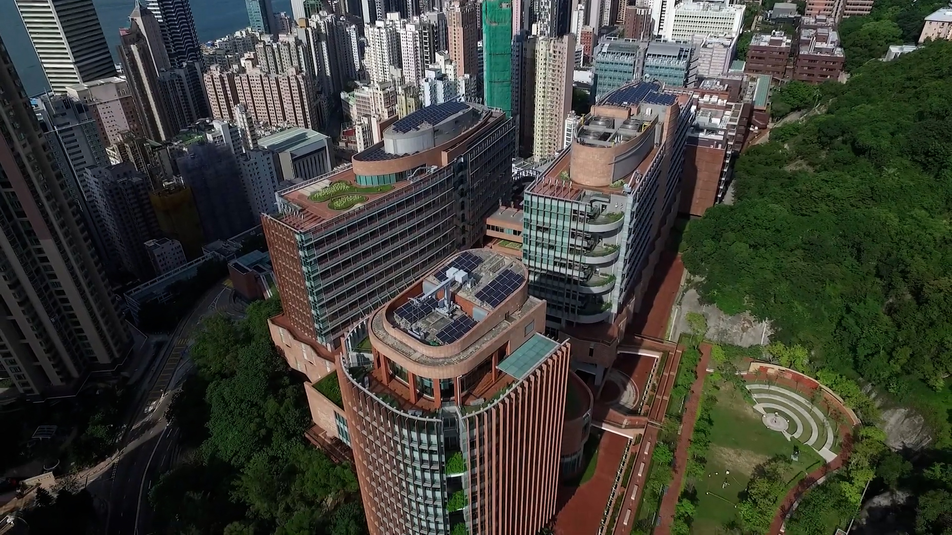 Aerial view of a modern urban campus with tall, curved buildings featuring rooftop gardens, surrounded by trees and city skyscrapers in the background.