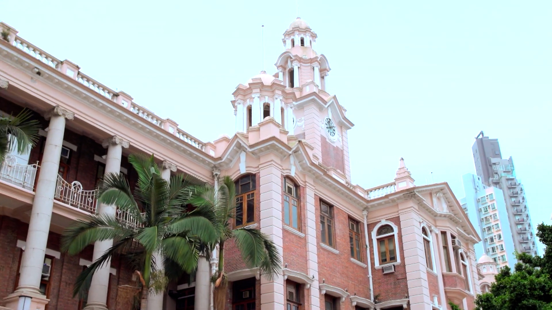 A historic red-brick building with white columns and a central clock tower, surrounded by palm trees, set against a clear sky with a modern skyscraper visible in the background.