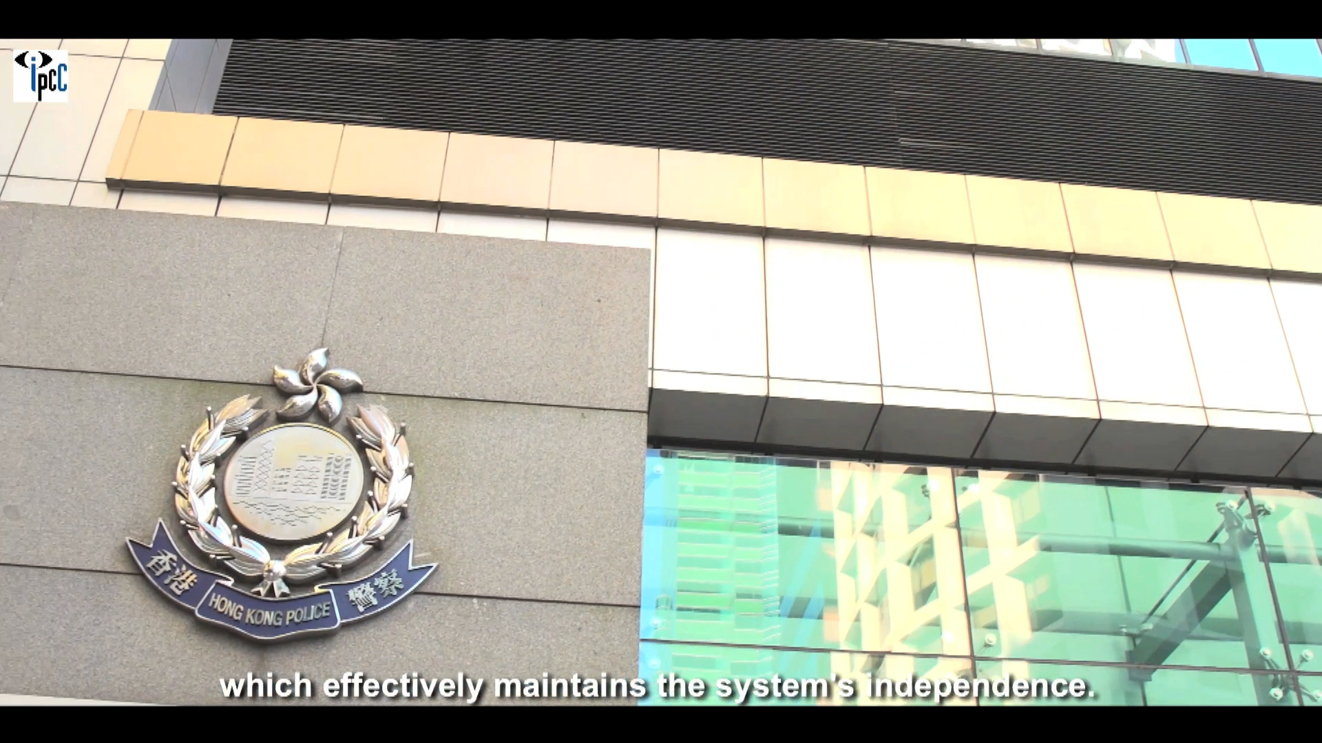 A building facade with the Hong Kong Police emblem mounted on the wall, next to a large window reflecting other buildings; partial subtitle at the bottom reads, "which effectively maintains the system's independence.
