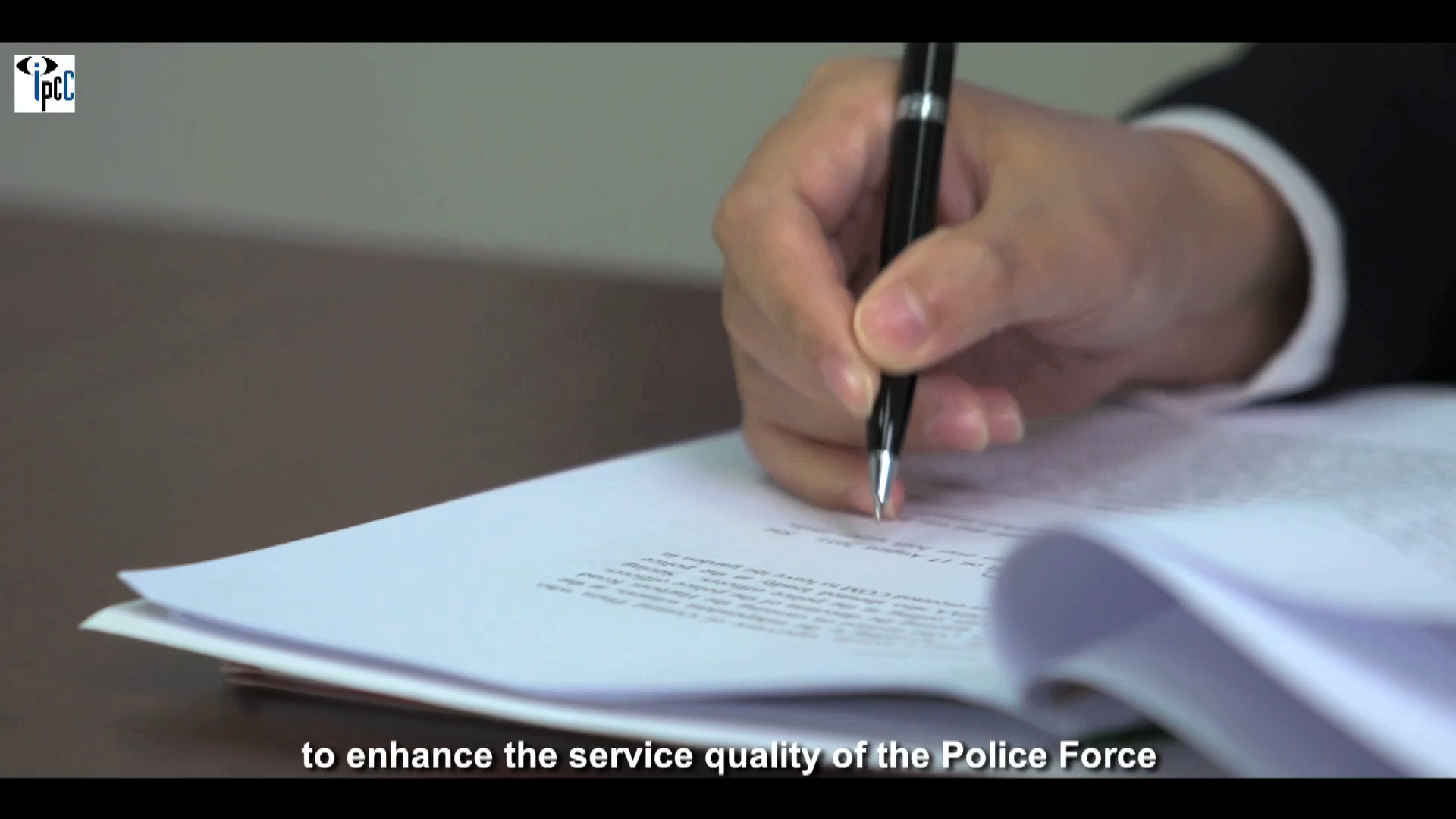 A close-up of a hand holding a pen and writing on a stack of papers. The text at the bottom reads, “to enhance the service quality of the Police Force.”.