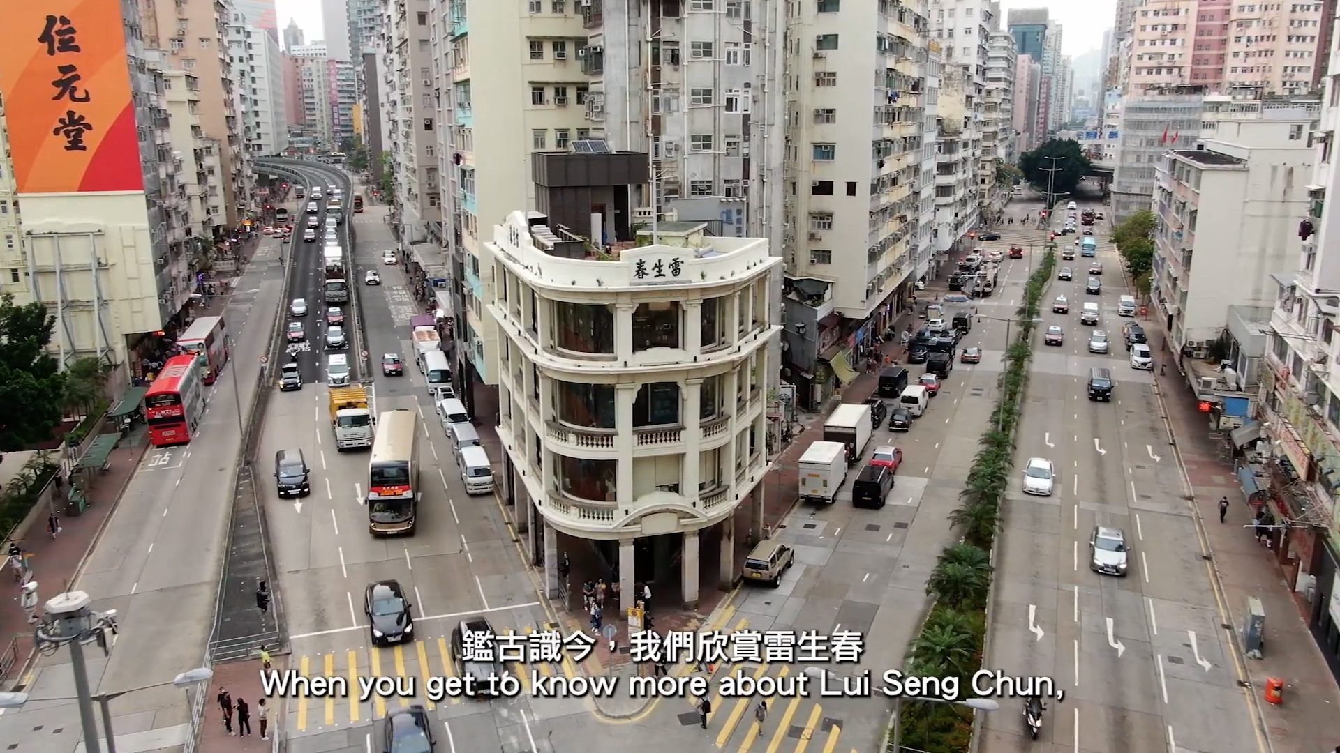 Aerial view of Lui Seng Chun, a historic white building with rounded corners situated at a busy intersection in an urban city filled with mid-rise apartments, traffic, and pedestrians. Subtitles in English and Chinese appear at the bottom.
