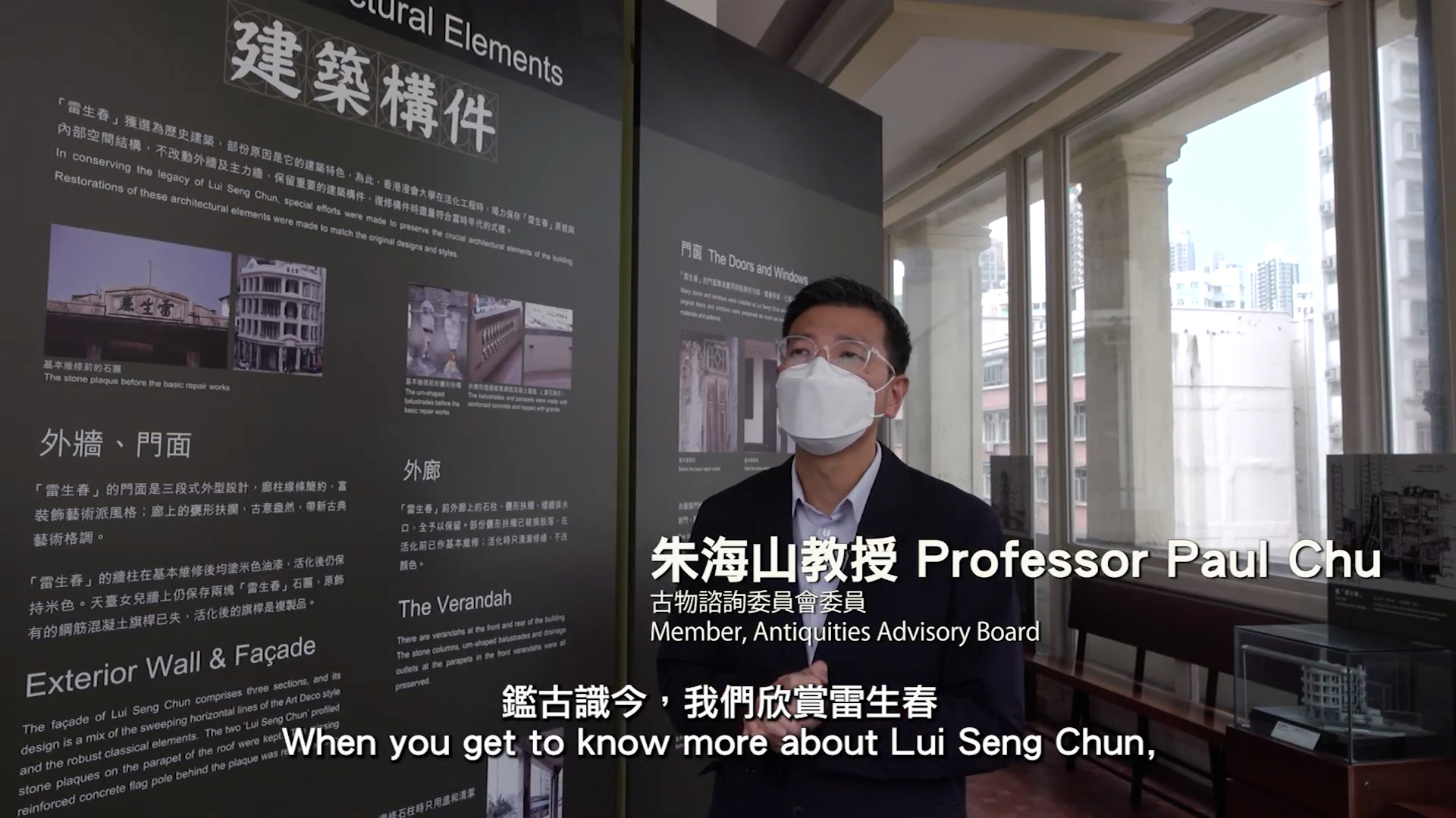 A man wearing glasses and a white mask stands indoors next to informational display boards in English and Chinese about architectural elements. Subtitles at the bottom read: "When you get to know more about Lui Seng Chun.