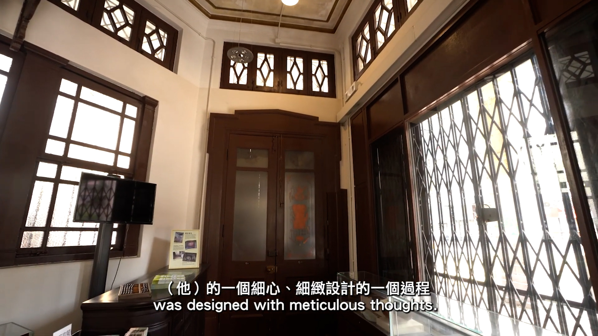 A small, sunlit room with large windows, wooden trim, a television on a table, informational brochures, and Chinese and English subtitles at the bottom of the image.
