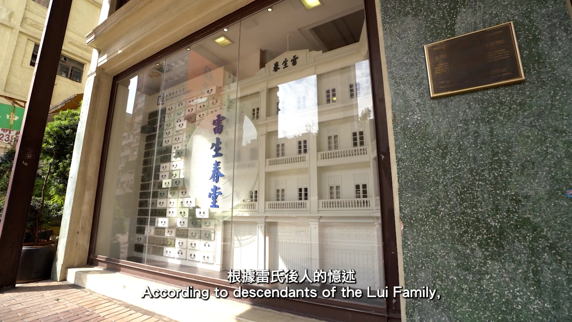 A storefront display window shows a detailed white model of a traditional building with Chinese characters above it. A plaque is mounted on the green wall to the right. English and Chinese subtitles appear at the bottom.