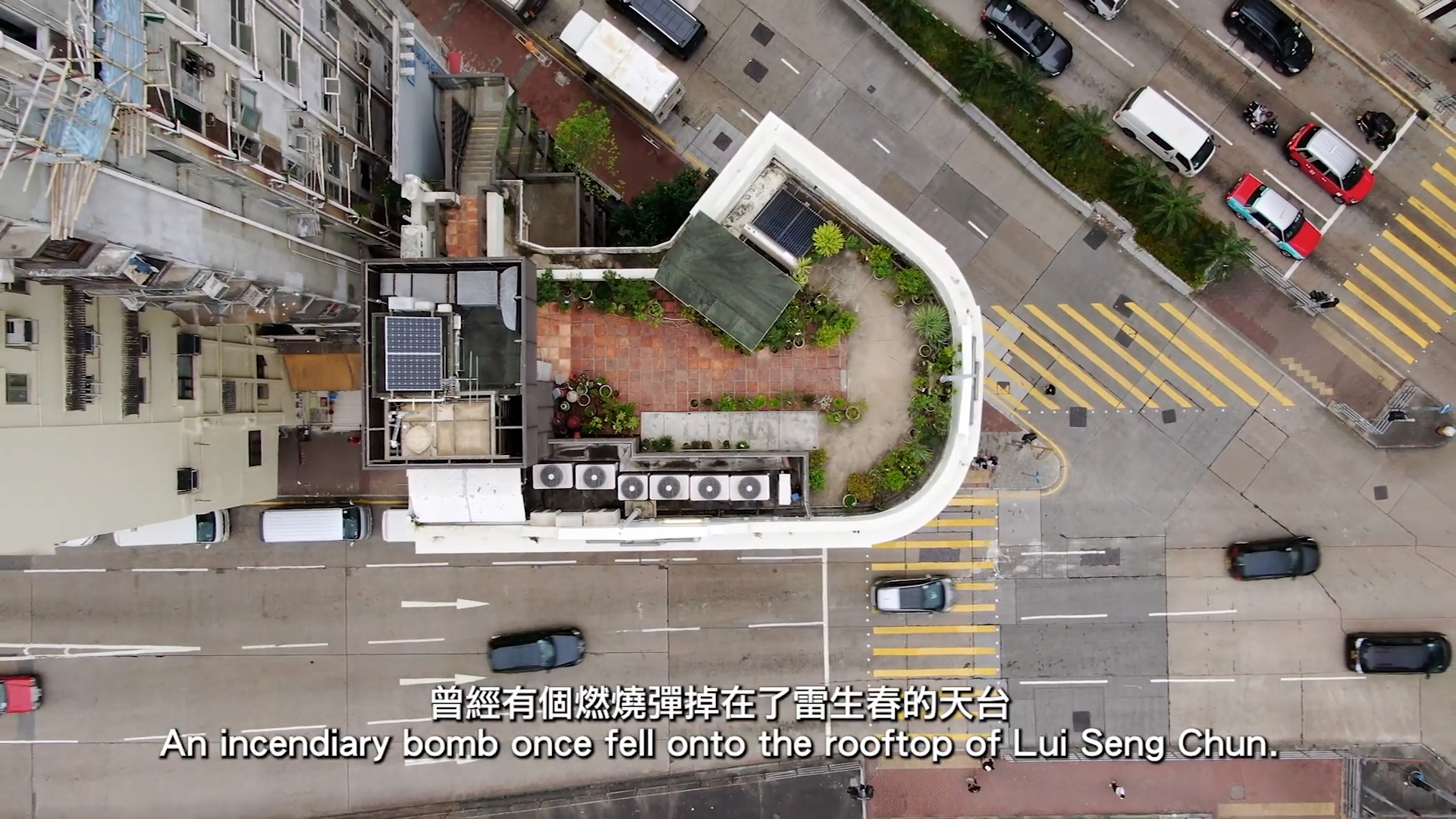 Aerial view of a city rooftop garden with potted plants, air conditioning units, and nearby roads; English and Chinese subtitles mention an incendiary bomb once fell on this rooftop.