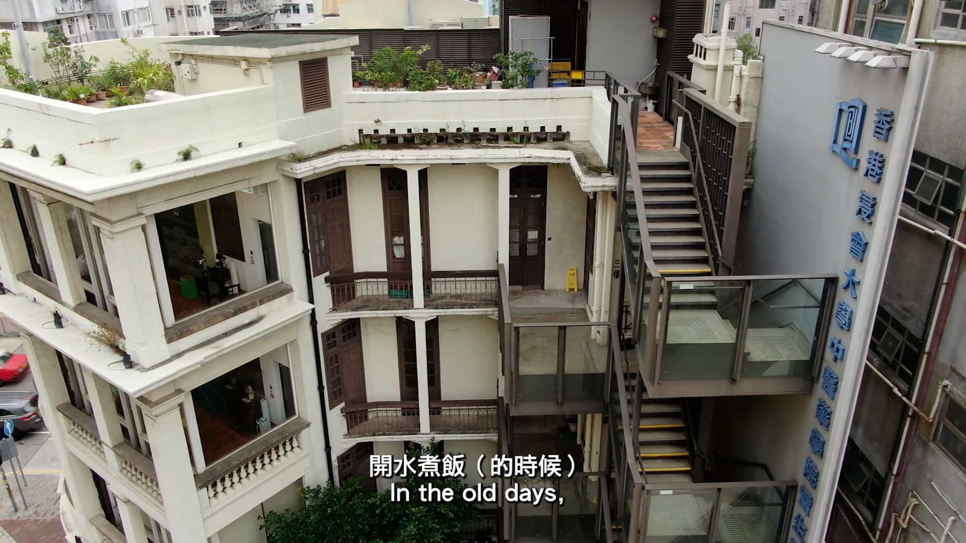 A multi-story building with rooftop gardens and external staircases stands next to a glass elevator. Chinese and English subtitles at the bottom read, “In the old days.”.