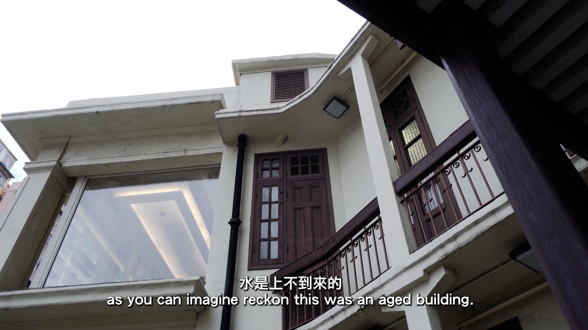 Low-angle view of an old, cream-colored building with wooden doors and windows, some glass panels, and subtitles at the bottom that read, “as you can imagine reckon this was an aged building.”.