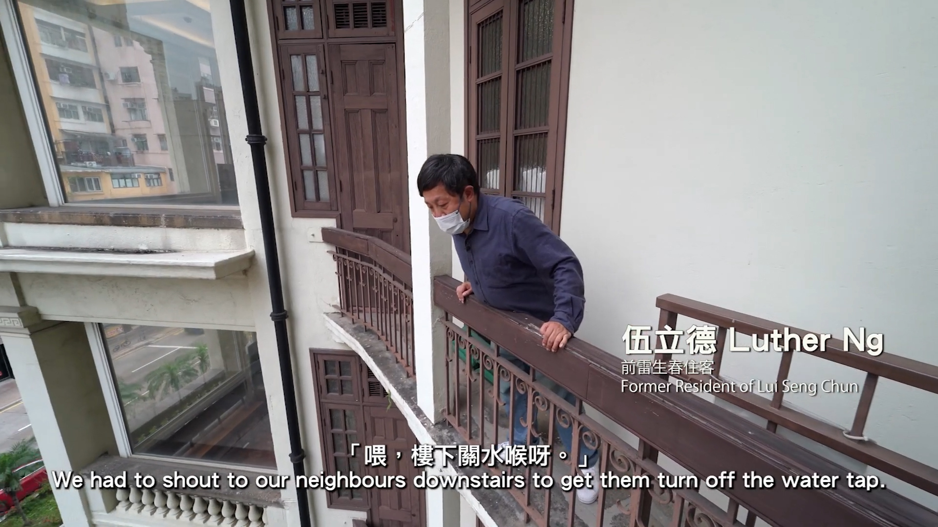 A man wearing a mask leans over a balcony railing of an old building. On-screen text reads: "Luther Ng, Former Resident of Lui Seng Chun." Subtitles say, "We had to shout to our neighbours downstairs to get them turn off the water tap.