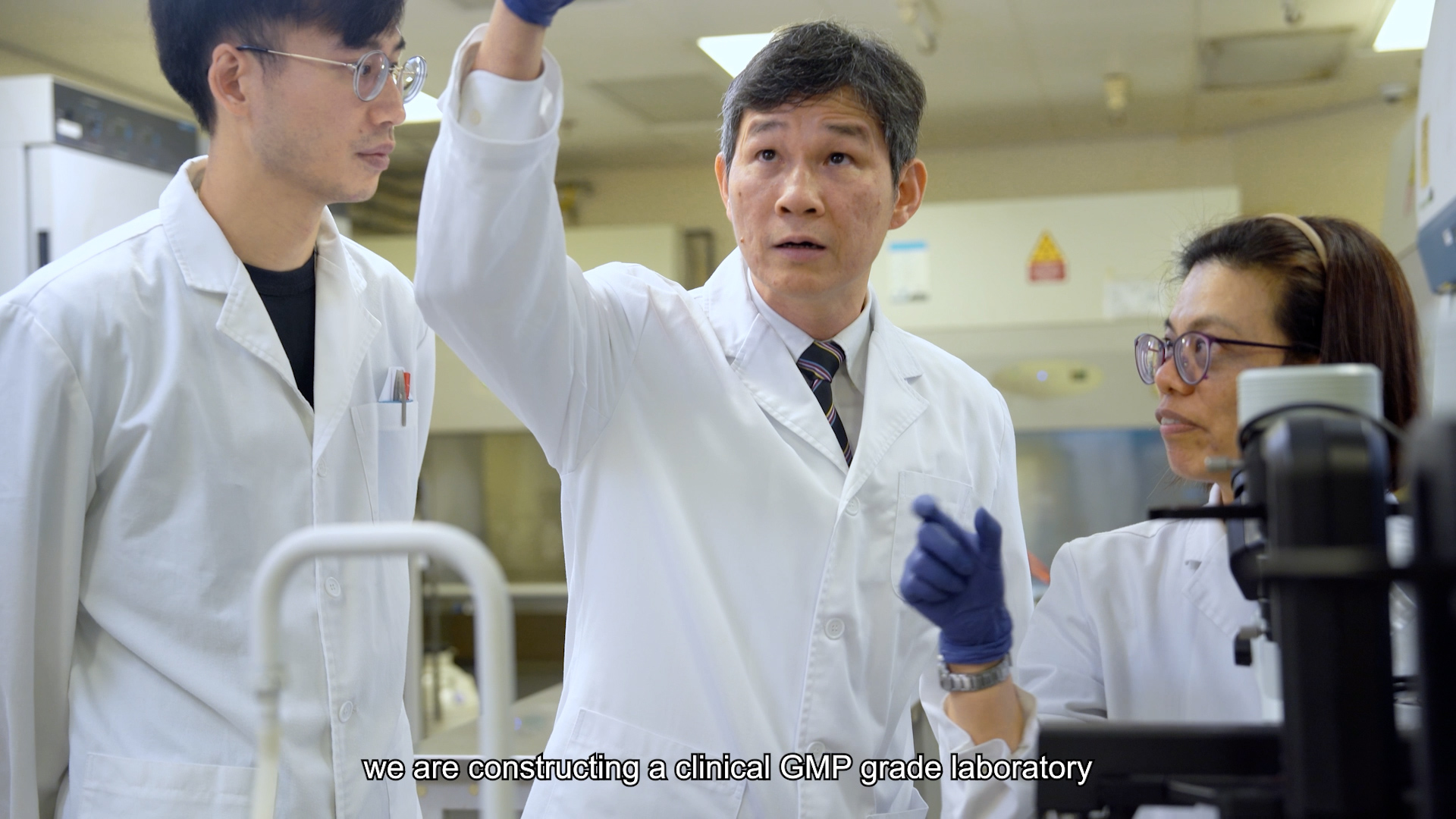 Three people in lab coats and gloves stand in a laboratory; the man in the center points and speaks while the others listen. Subtitle reads: "we are constructing a clinical GMP grade laboratory.
