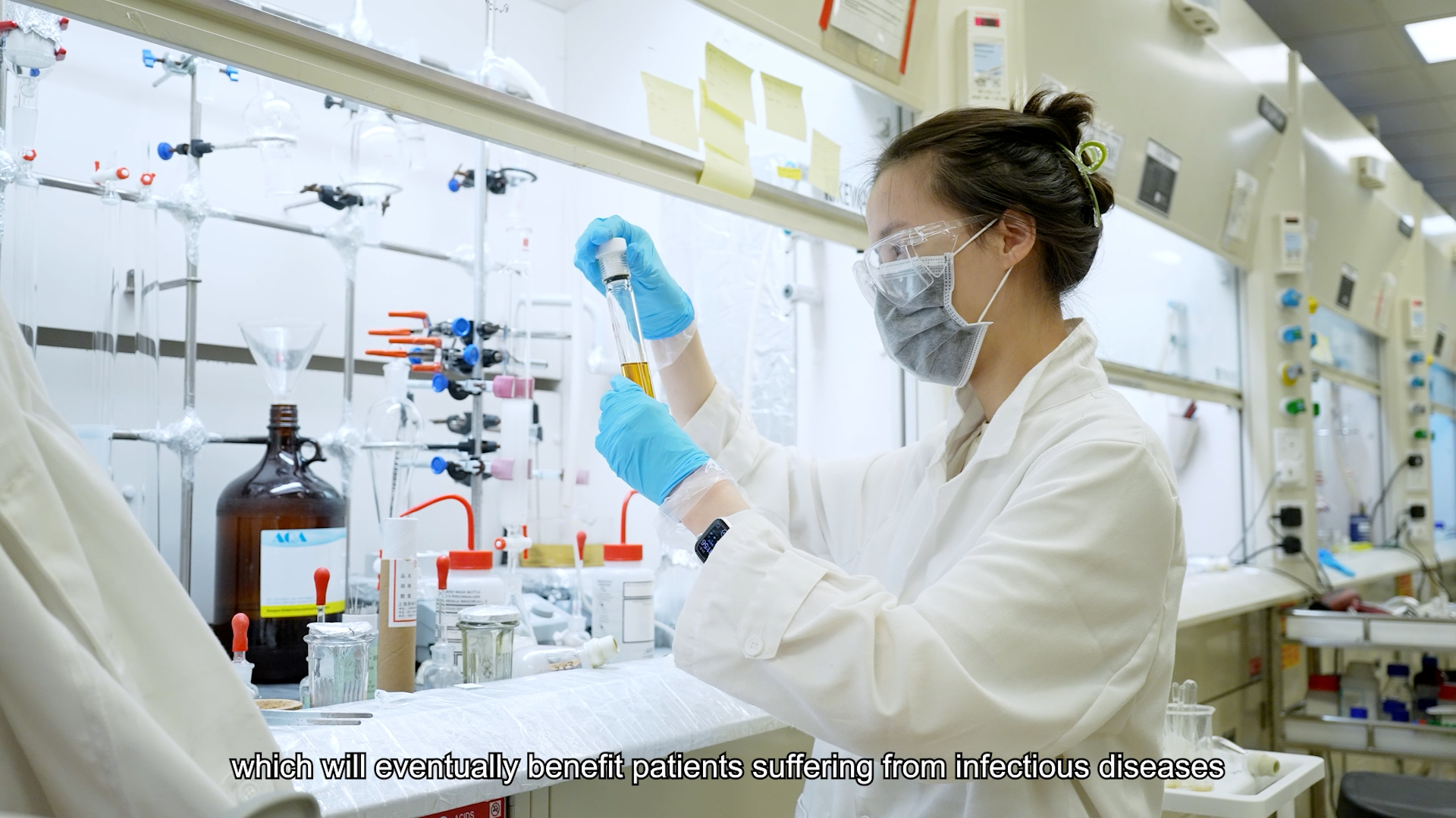 A scientist wearing a lab coat, gloves, mask, and goggles works with lab equipment and test tubes in a laboratory. Subtitle reads: "which will eventually benefit patients suffering from infectious diseases.