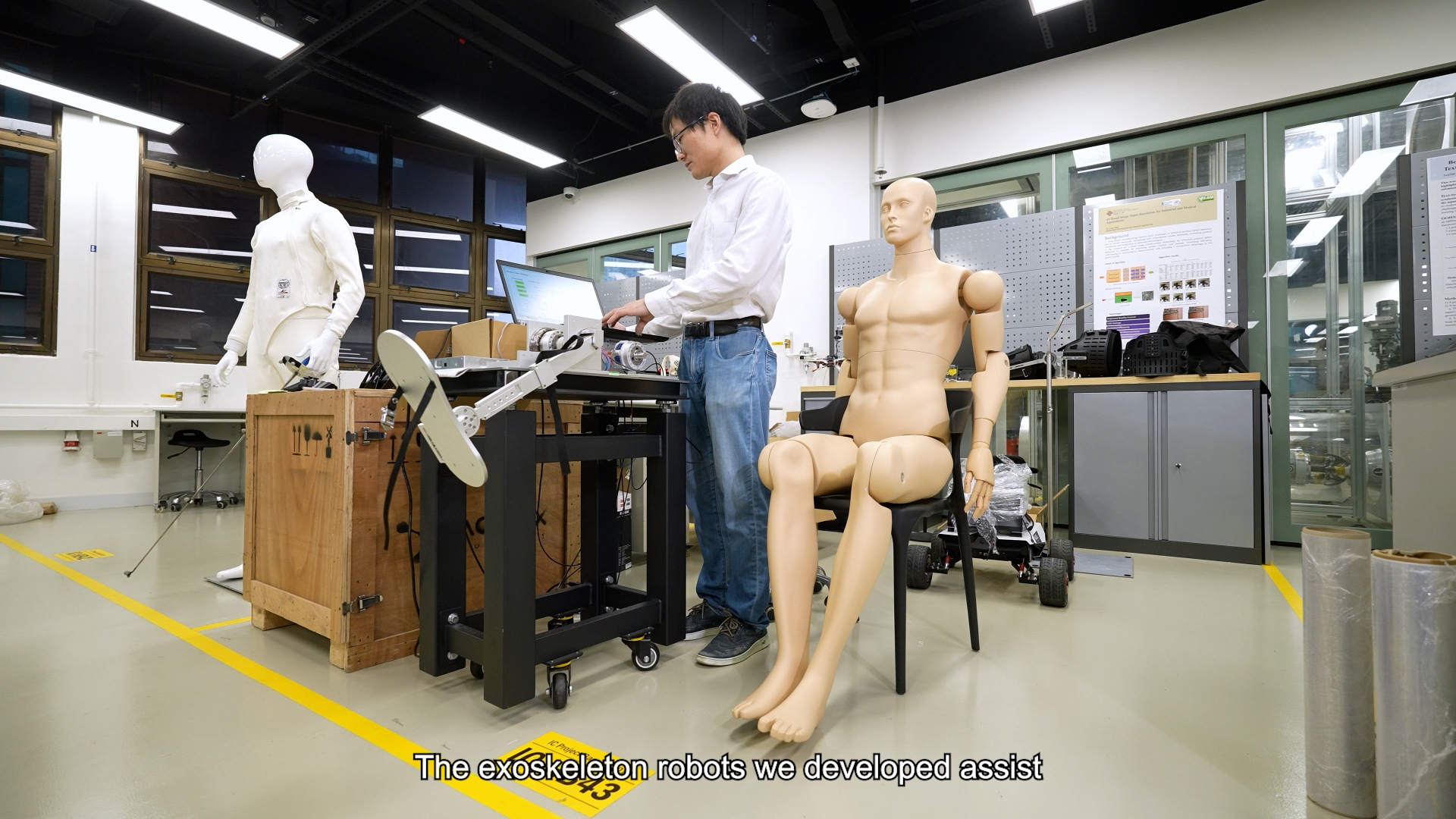 A person tests an exoskeleton device on a seated mannequin in a laboratory, with another mannequin and technical equipment visible. Subtitle reads, "The exoskeleton robots we developed assist.