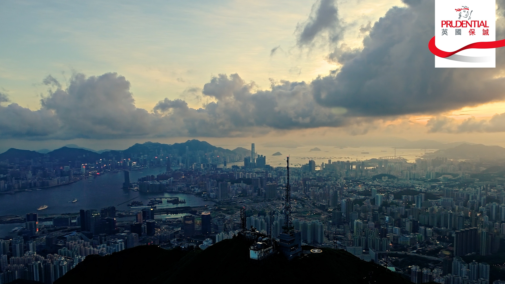 Aerial view of Hong Kong at sunset with dramatic clouds, city skyline, and harbor. The Prudential logo appears in the top right corner.