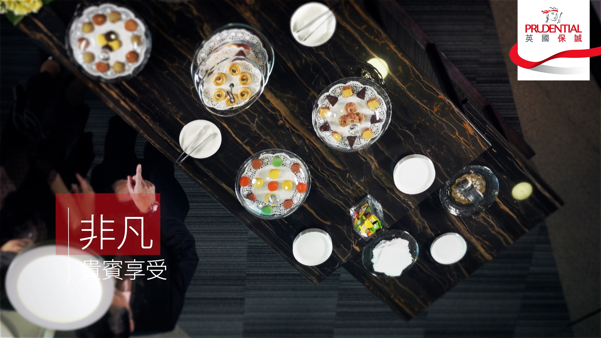 Overhead view of a dark marble table set with various desserts, drinks, and empty bowls. Two people in black attire are seated at the table. Prudential logo and Chinese text are visible in the top right and bottom left corners.