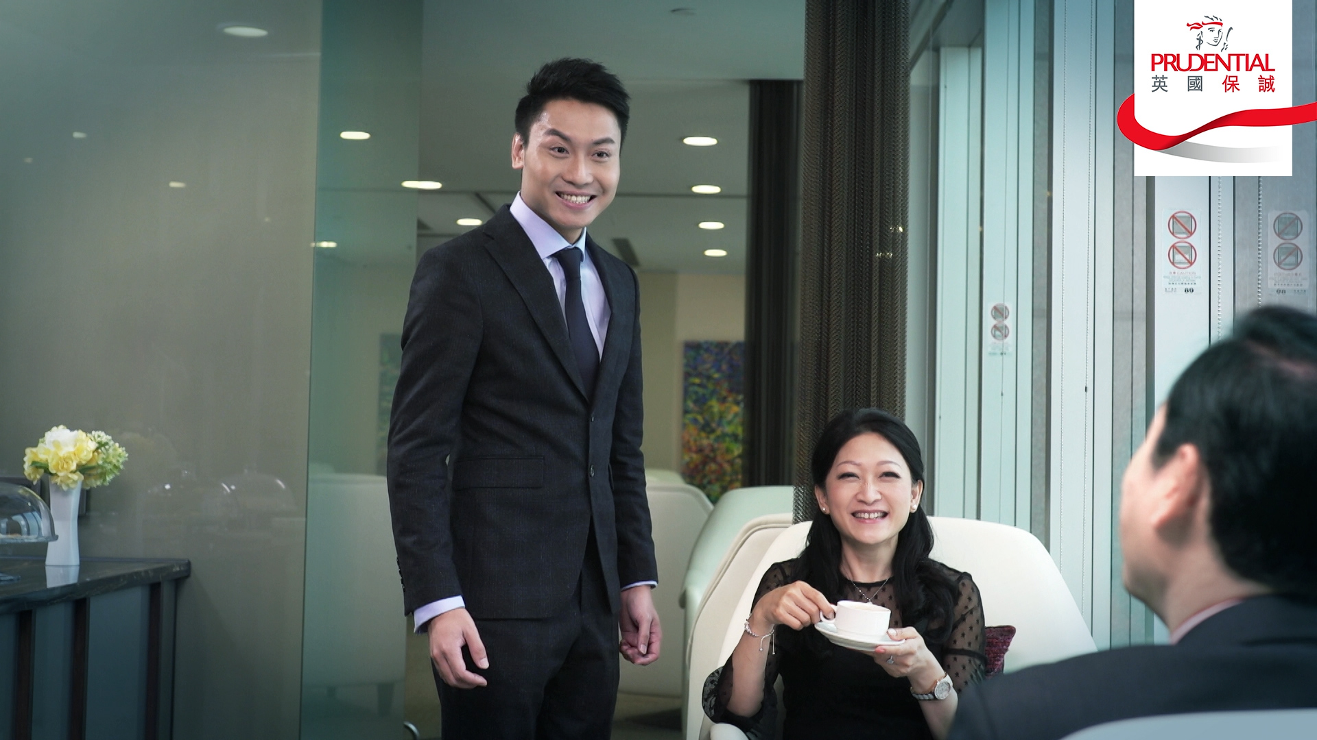 A man in a dark suit stands and smiles next to a seated woman holding a teacup, both interacting with another seated man. The setting appears to be an office lounge with Prudential branding visible.