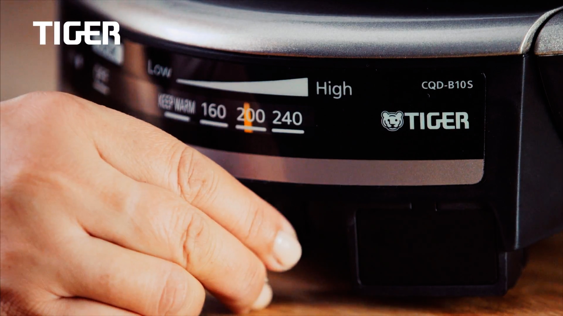 A hand adjusts the heat setting dial on a TIGER brand appliance, with the temperature range showing from low to high, and the pointer aligned at 200°C.