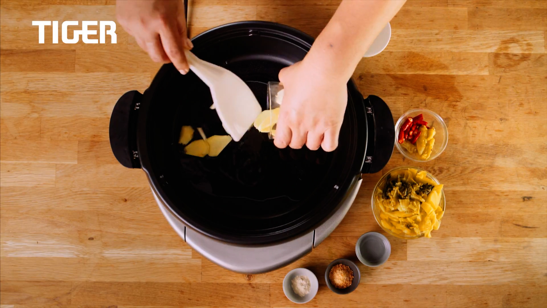 Top-down view of hands adding sliced ginger and butter into a black pan. Surrounding the pan are small bowls containing various ingredients like spices, chilies, and pickled vegetables. "TIGER" logo is in the corner.