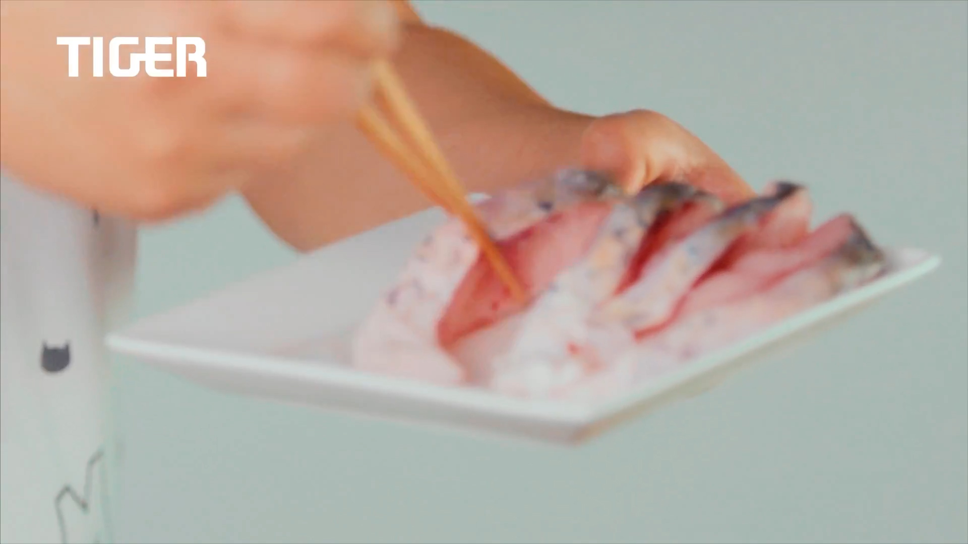 A person uses chopsticks to pick up a piece of sashimi from a white plate holding several slices of raw fish. The word "TIGER" appears in white text in the top left corner.