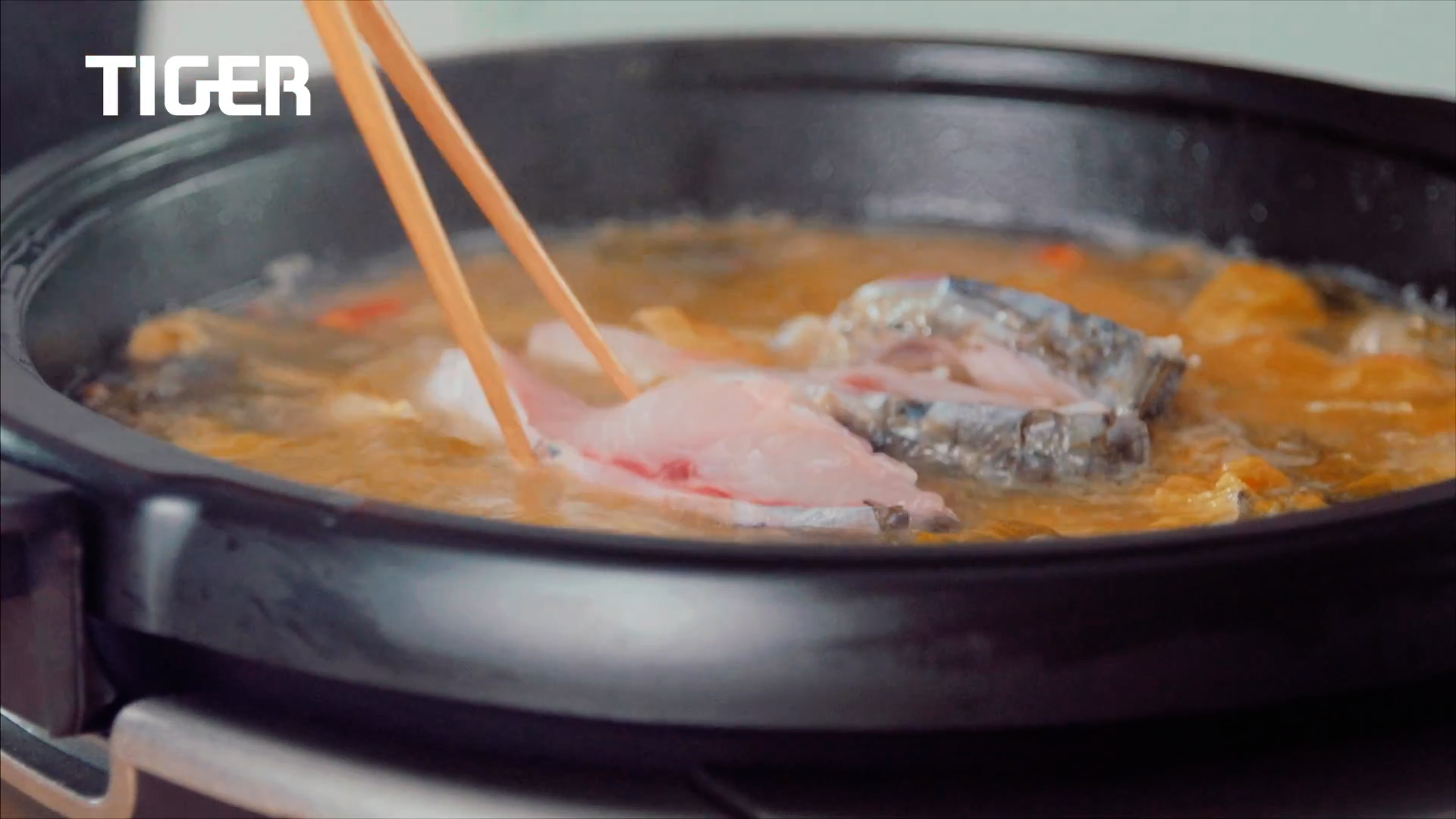 A close-up of chopsticks picking up a piece of fish from a simmering pot of soup, with fish and vegetables visible in the broth. The word "TIGER" appears in the upper left corner.
