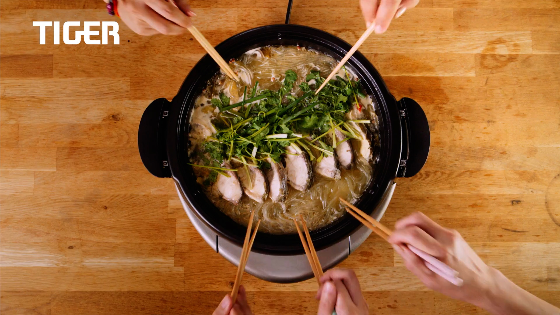 Four pairs of hands using chopsticks to pick food from a hot pot filled with noodles, herbs, and sliced meat, placed on a wooden table. The word "TIGER" appears in the top left corner.