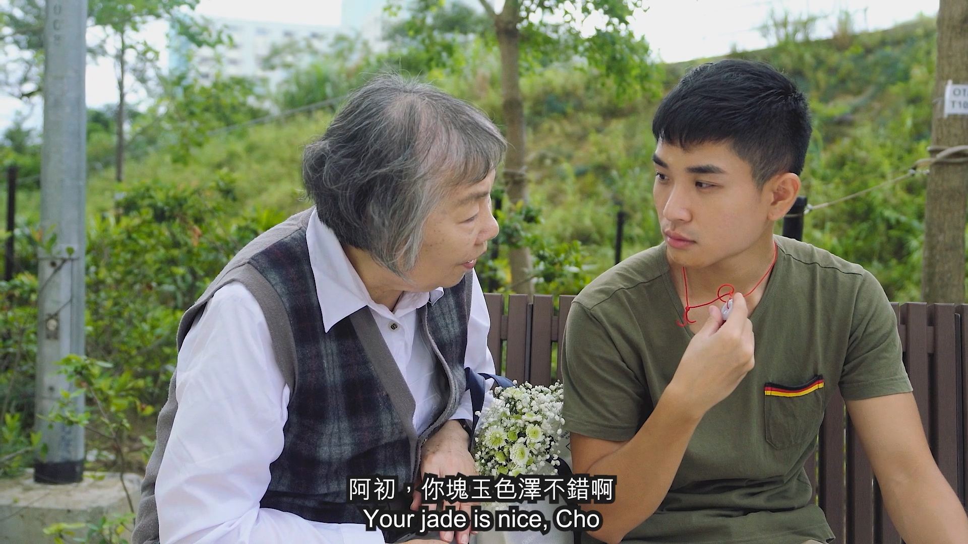 An elderly woman and a young man sit on a bench outdoors. The woman, holding flowers, speaks to the man, who is showing her a jade pendant. English subtitles read, “Your jade is nice, Cho.” Mandarin text appears above the subtitles.