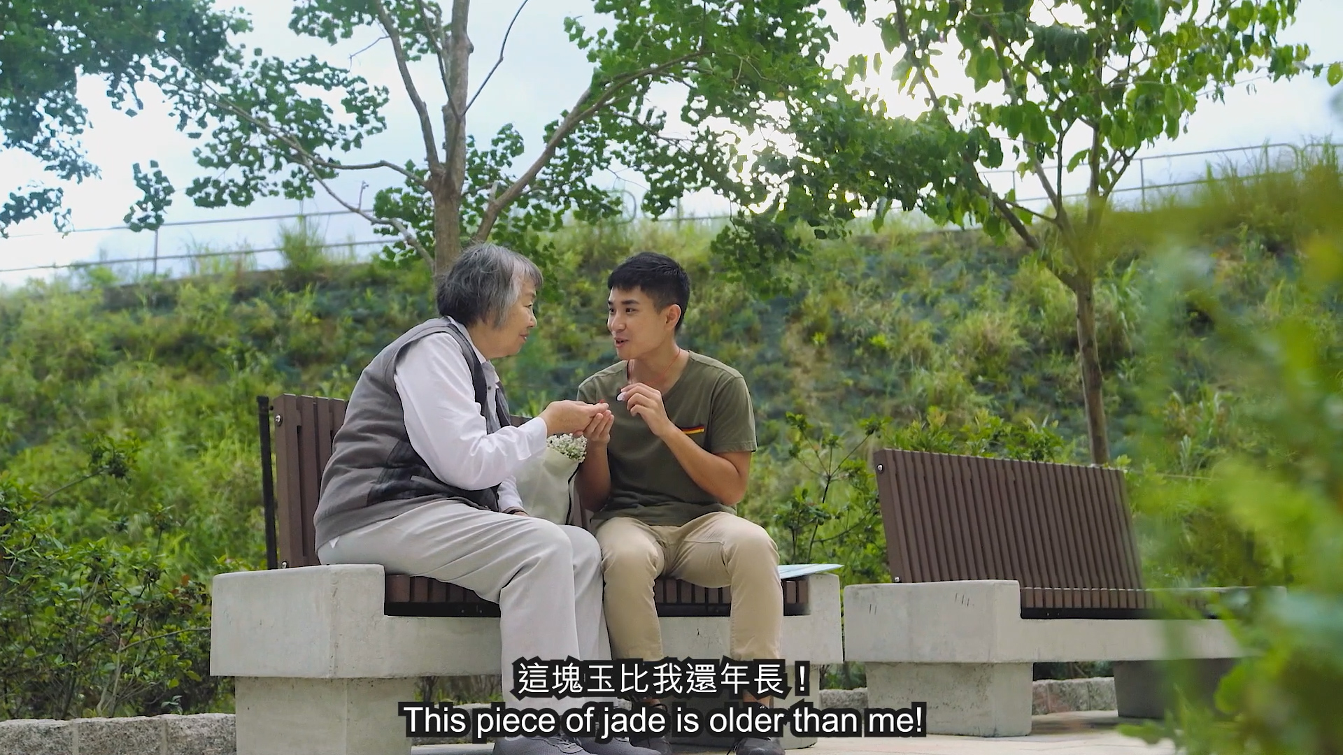 An elderly woman and a young man sit on a bench outdoors, talking and holding a jade object. Green trees and bushes are in the background. Subtitles read: "This piece of jade is older than me!.