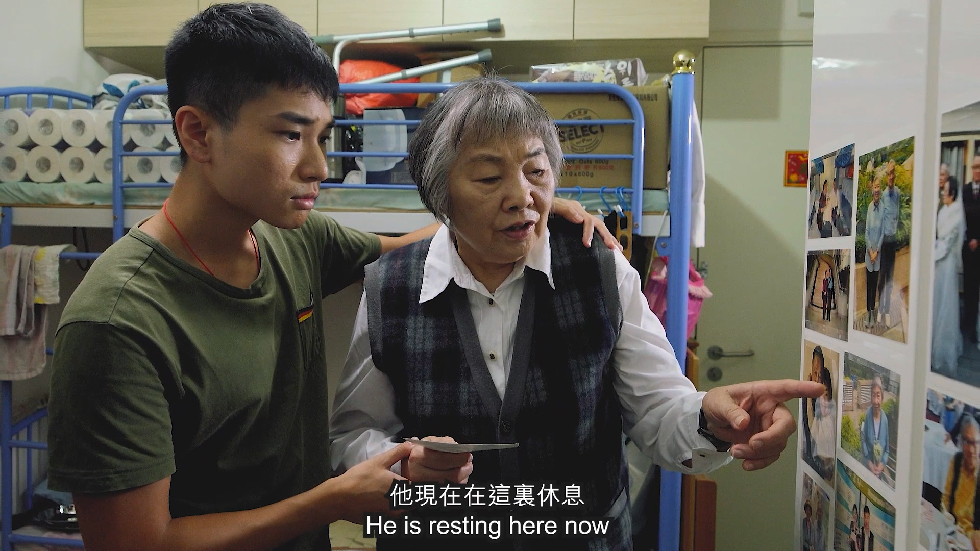A young man and an older woman stand together in a room filled with stacked toilet paper rolls. The woman points at photos on the wall, while the man holds a mug. English and Chinese subtitles read, "He is resting here now.