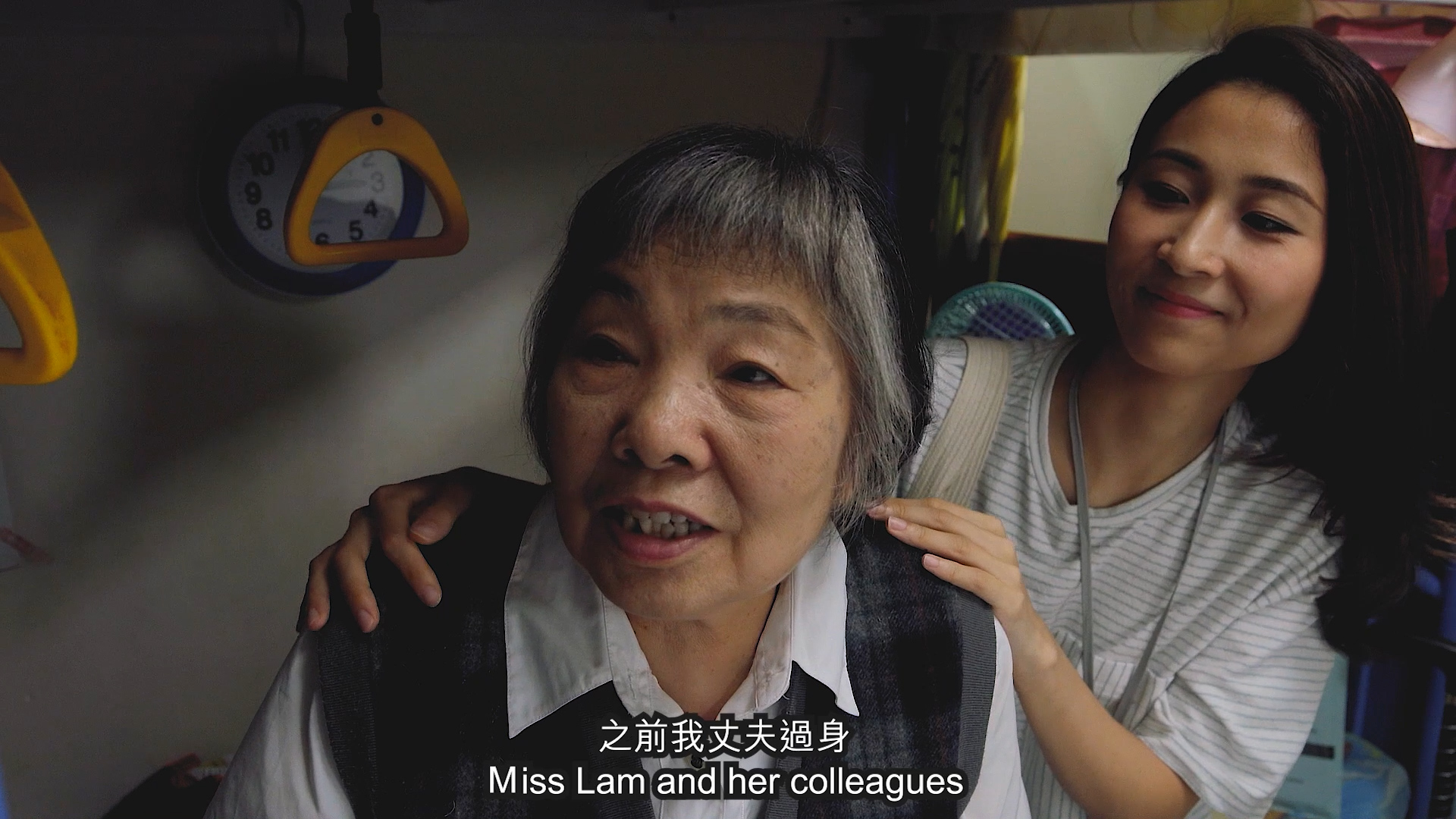 An older woman sits and smiles while a younger woman stands behind her, gently resting her hands on the older woman's shoulders. Subtitles read, "Miss Lam and her colleagues." Chinese text appears above the subtitles.