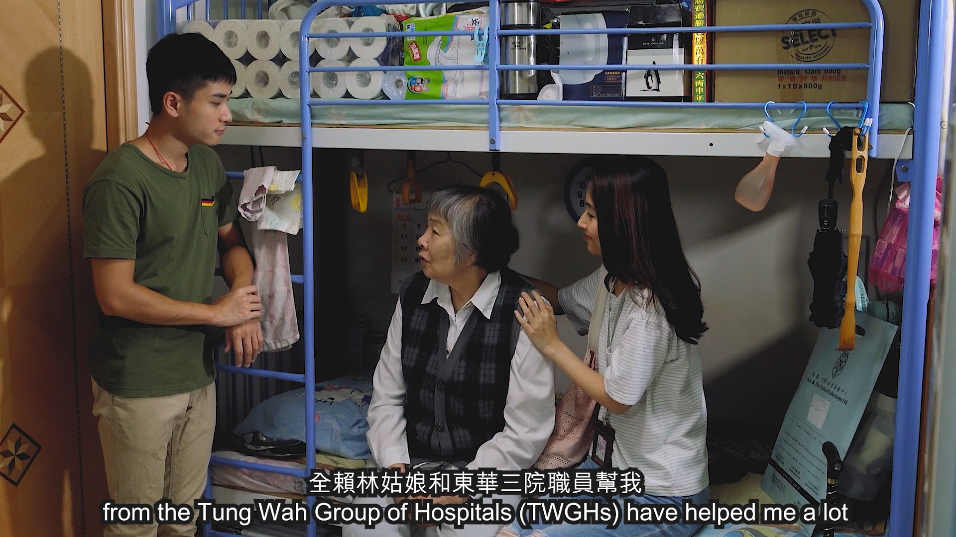 A young man speaks to an elderly woman sitting on a lower bunk bed, while a young woman sits beside her with a comforting hand on her back. The room is small with shelves stacked with household supplies. Subtitles are present.