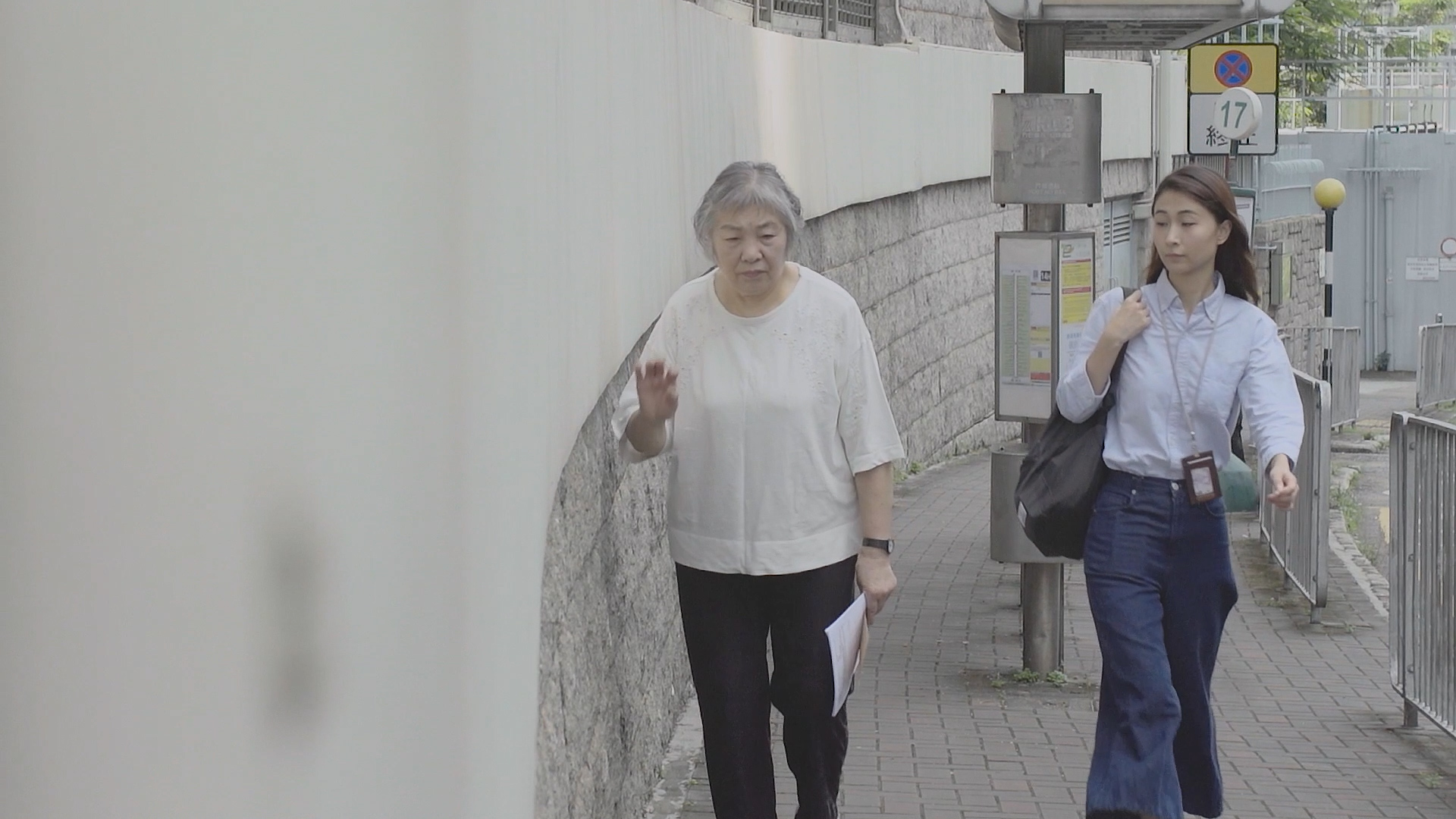 Two women walk on a city sidewalk beside a curved white wall; one is older with gray hair holding papers, the other is younger with long dark hair, carrying a shoulder bag and a phone clipped to her belt.