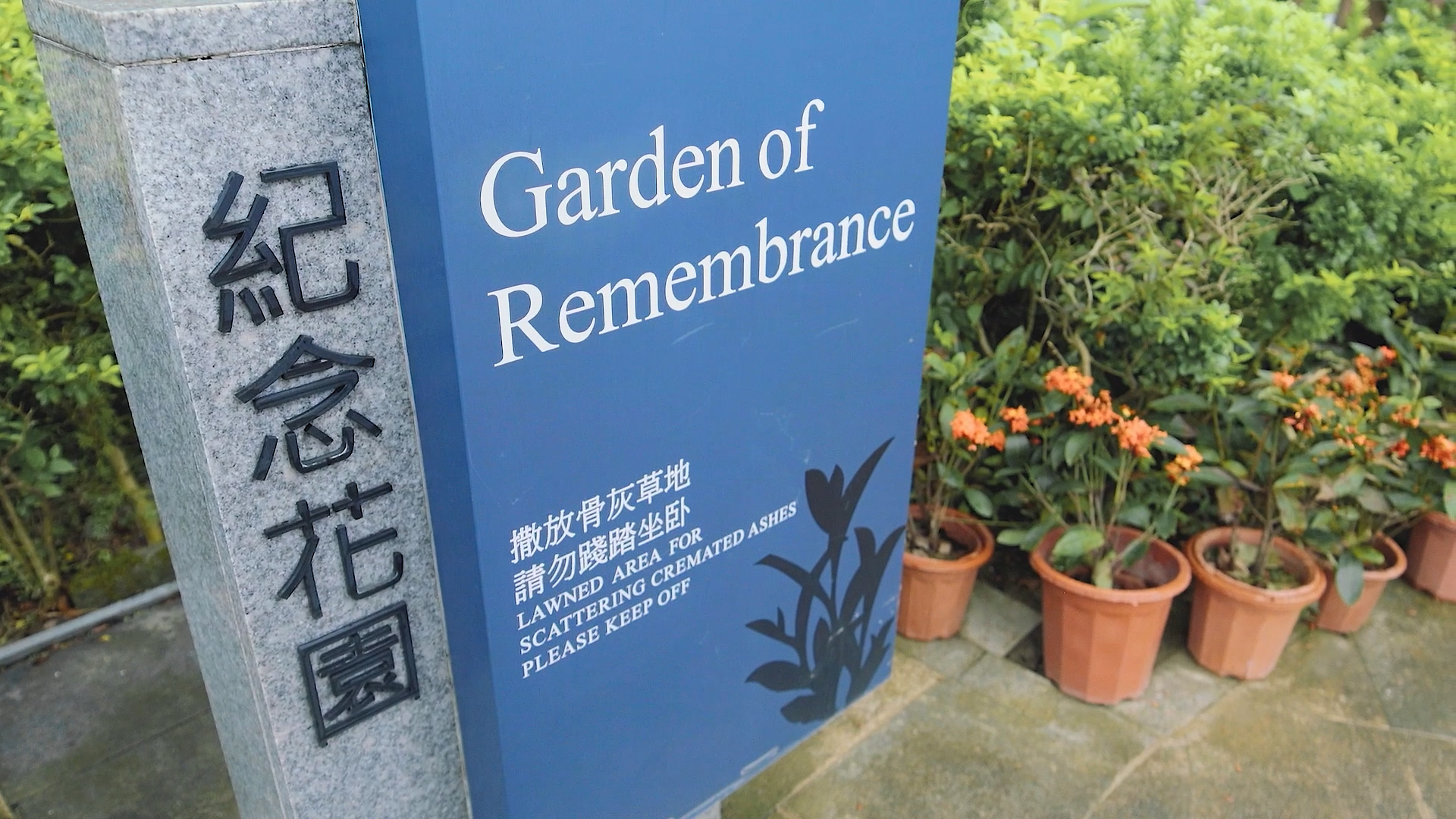 A blue sign reading "Garden of Remembrance" with Chinese text stands beside orange potted flowers and a stone pillar engraved with more Chinese characters in an outdoor garden setting.