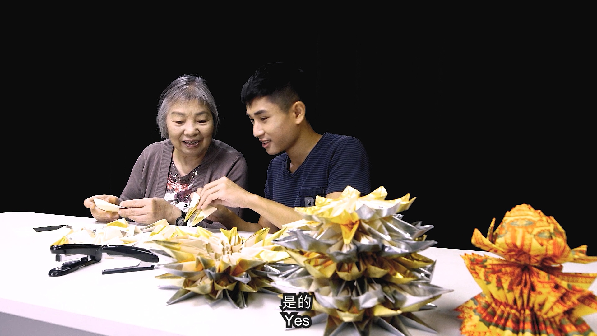 An older woman and a young man sit at a table, smiling and folding colorful origami. Several completed origami sculptures are on the table, and the caption "Yes" appears at the bottom of the image. The background is black.