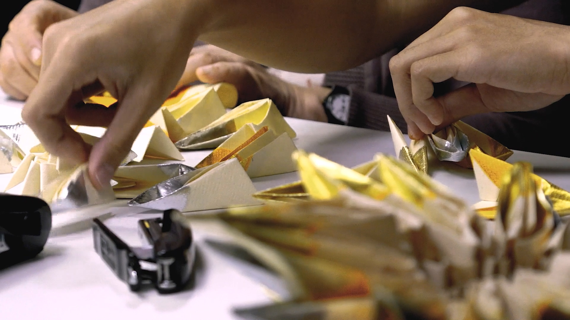 Close-up of hands folding gold, silver, and white origami paper on a table, with completed pieces nearby. A black stapler is also visible in the foreground.