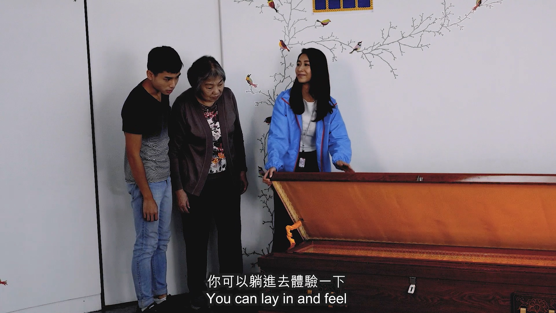 Three people stand beside an open, empty coffin in a room with bird and tree wall art. One person gestures toward the coffin. Subtitles read: "You can lay in and feel" in English and Chinese.