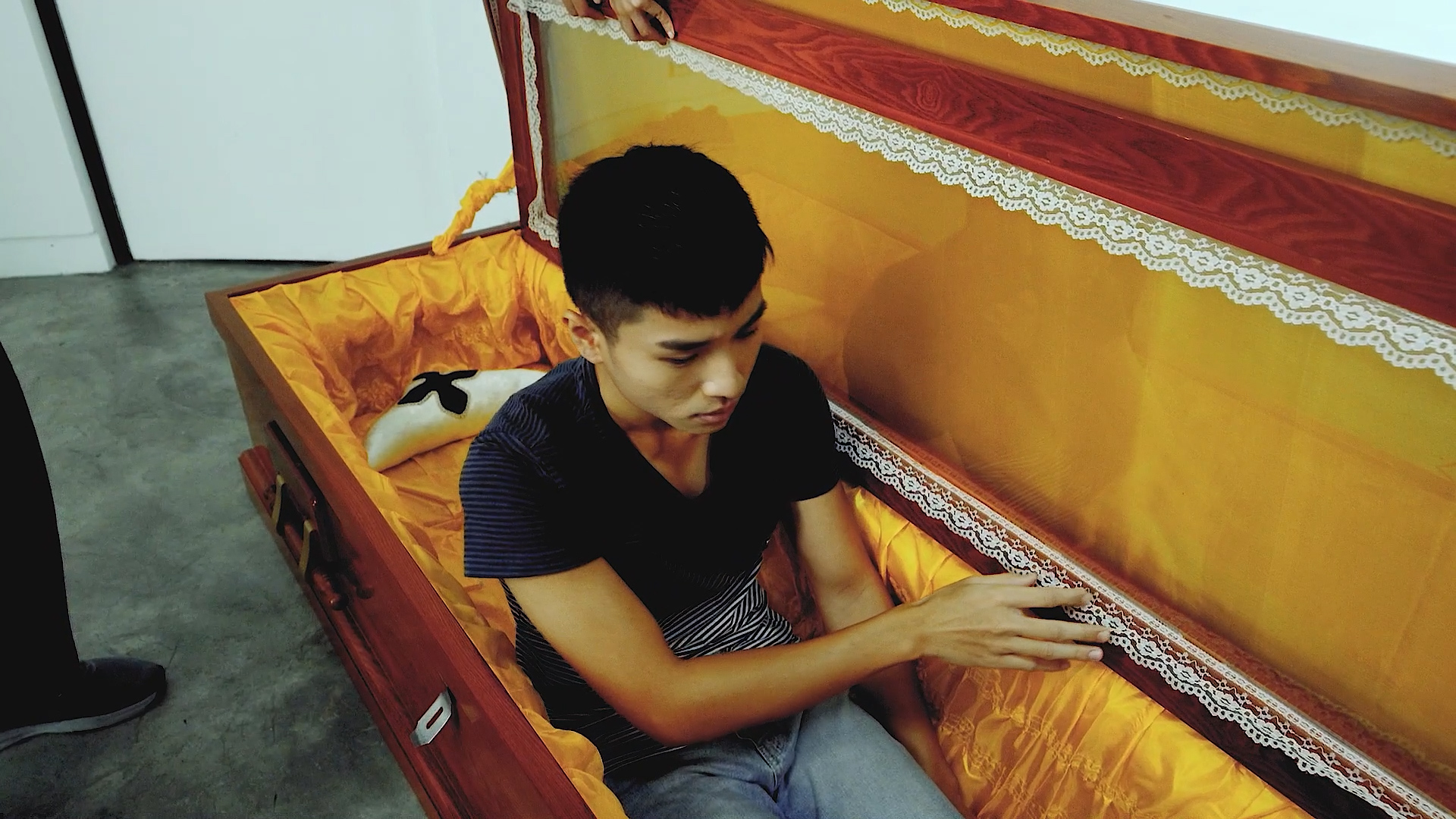 A young man sits inside an open, orange-lined coffin, looking down while touching the side. Another person’s hand is visible holding the lid. The scene appears to be indoors on a grey floor.