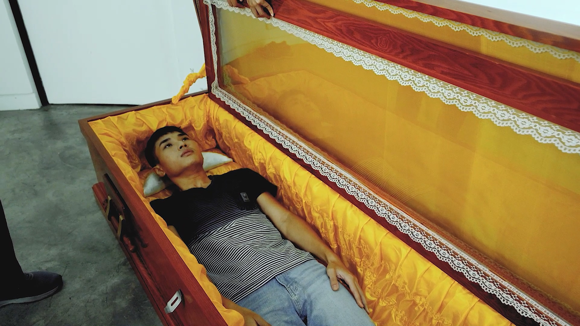A young man lies inside an open coffin lined with yellow fabric, wearing a black and white striped shirt and jeans. Someone's hand is holding the coffin lid, appearing to close or open it.