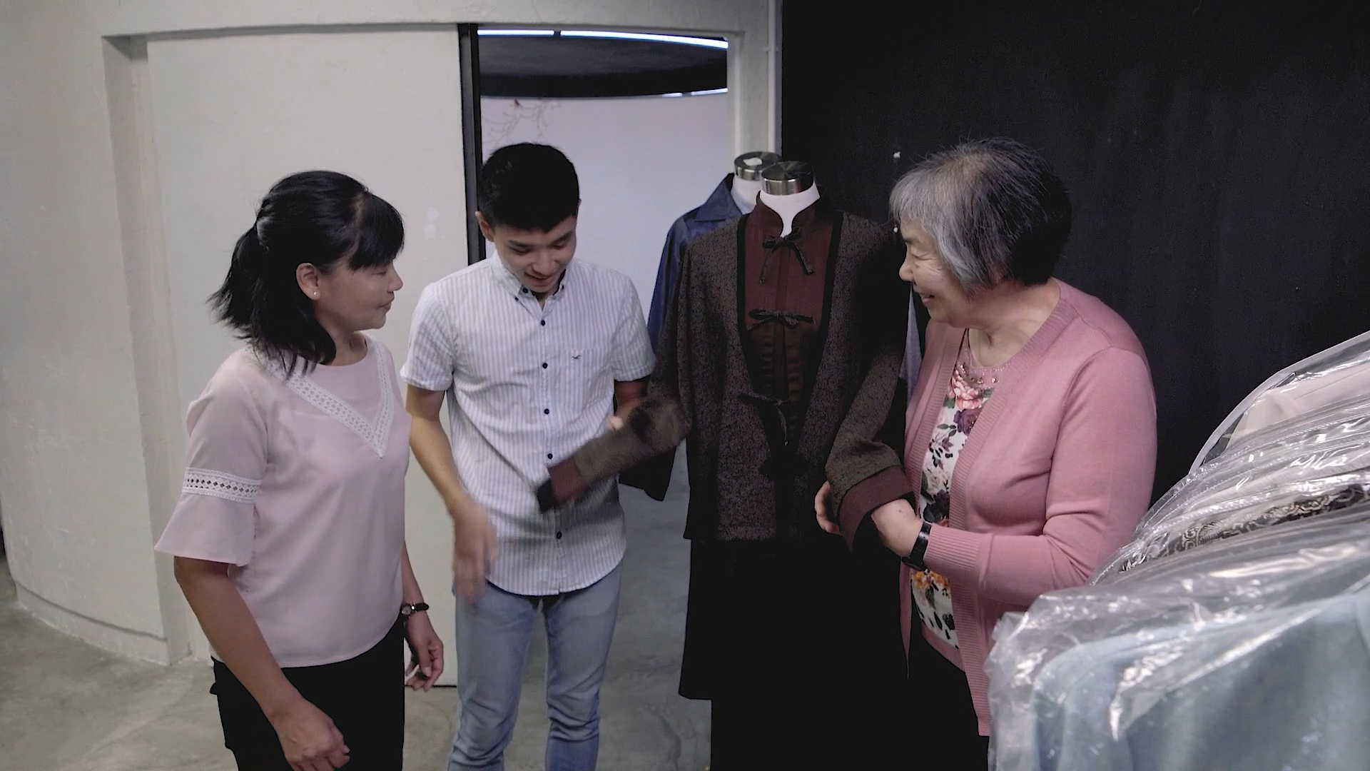 Three people stand near a clothing rack and a mannequin wearing a brown outfit. The older woman in pink holds the mannequin’s sleeve while the younger man and woman look and converse in a well-lit room.