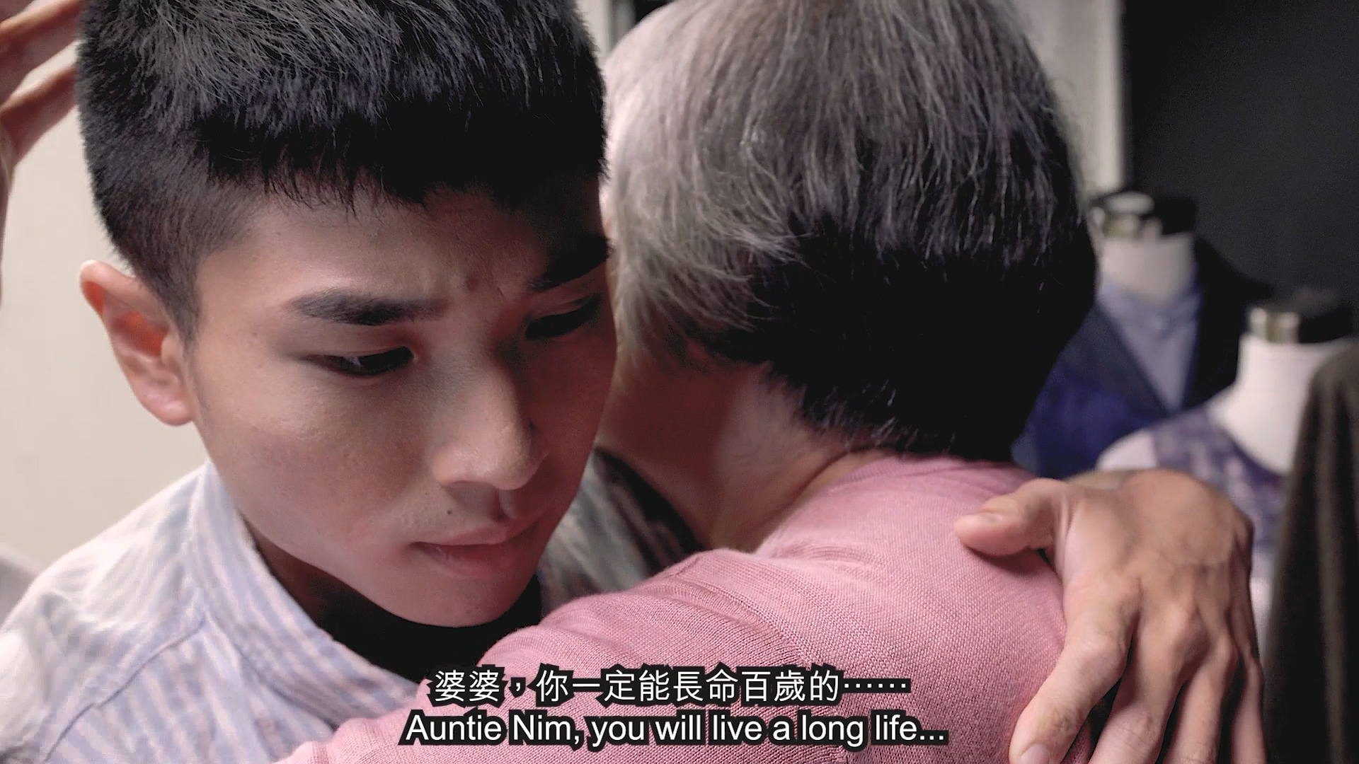 A young man hugs an older woman with gray hair, looking serious. The woman wears a pink top. The subtitles read: "Auntie Nim, you will live a long life...