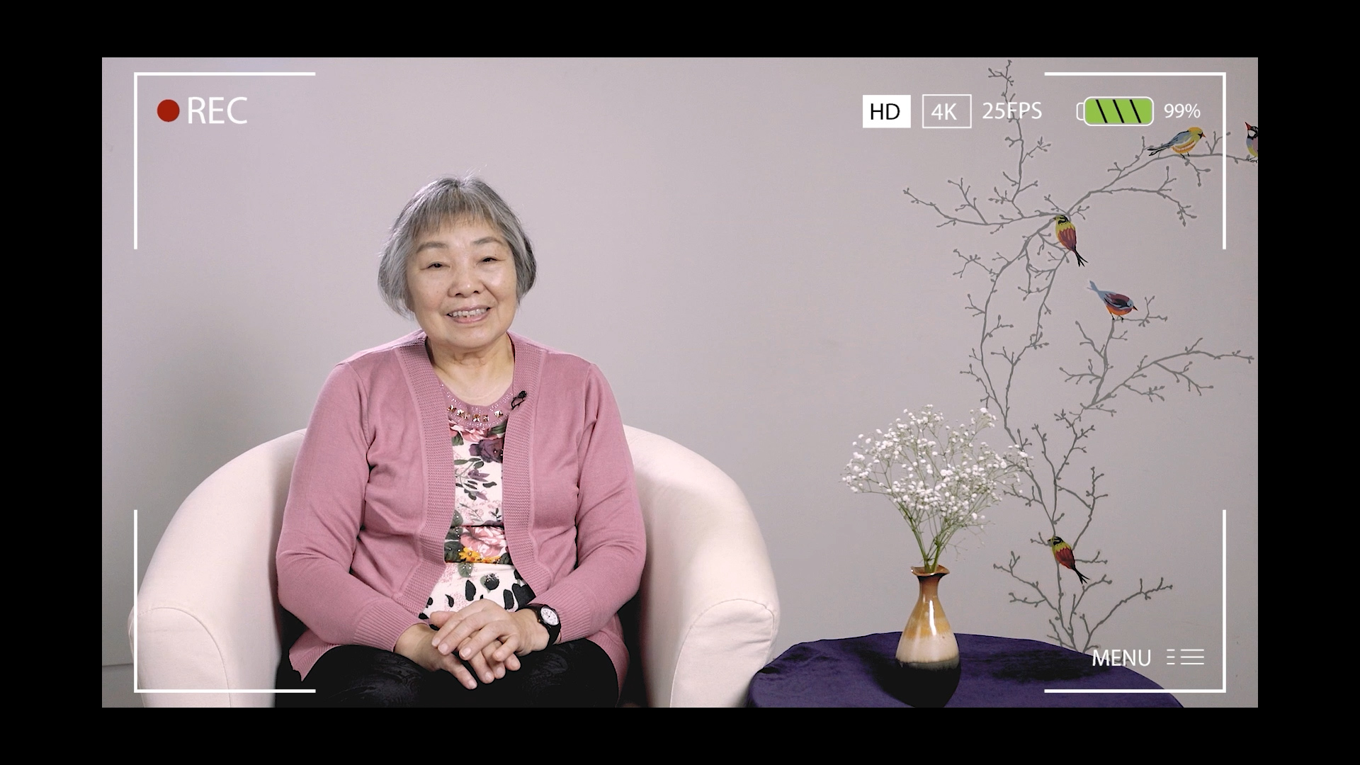 An older woman sits in a white chair, smiling, wearing a pink cardigan and floral top. Behind her is a wall with a bird and tree branch design, and a vase with white flowers on a small table. Camera recording graphics are visible.
