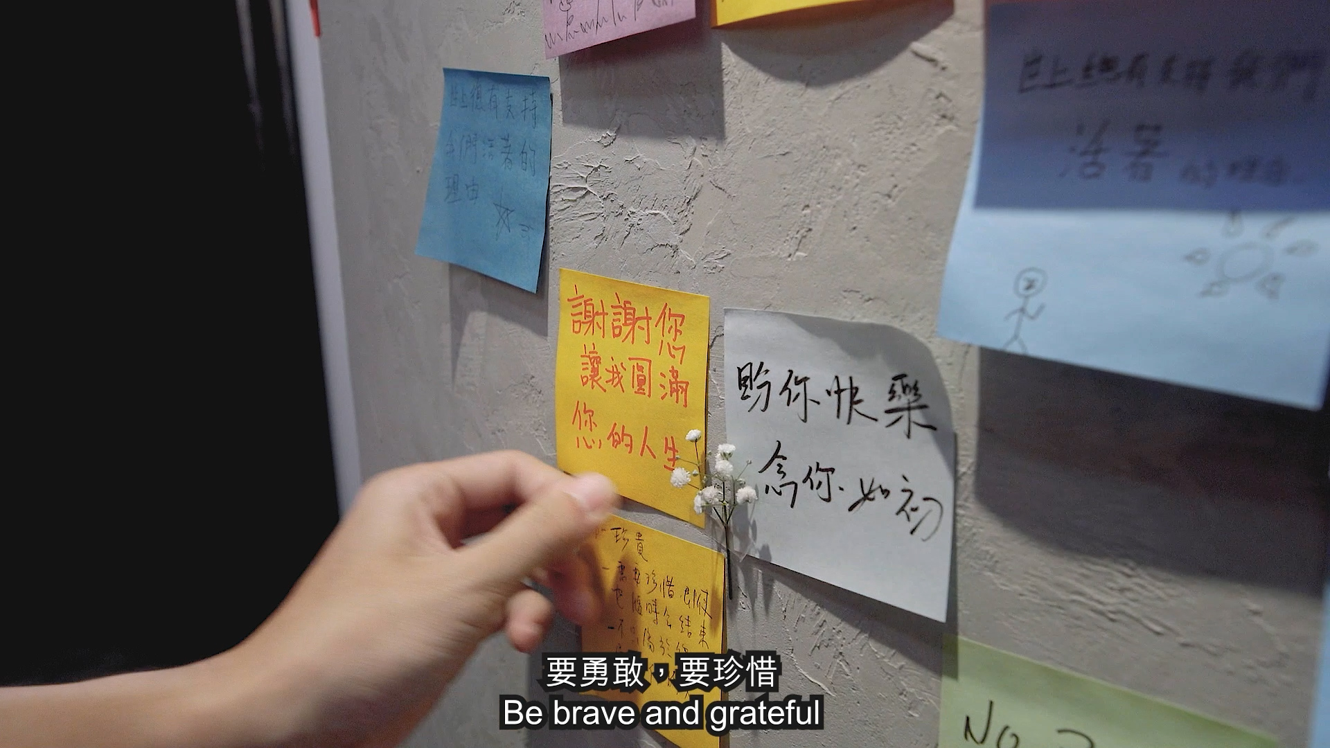 A hand pins colorful sticky notes with Chinese writing onto a textured wall. Subtitles at the bottom read, "Be brave and grateful.