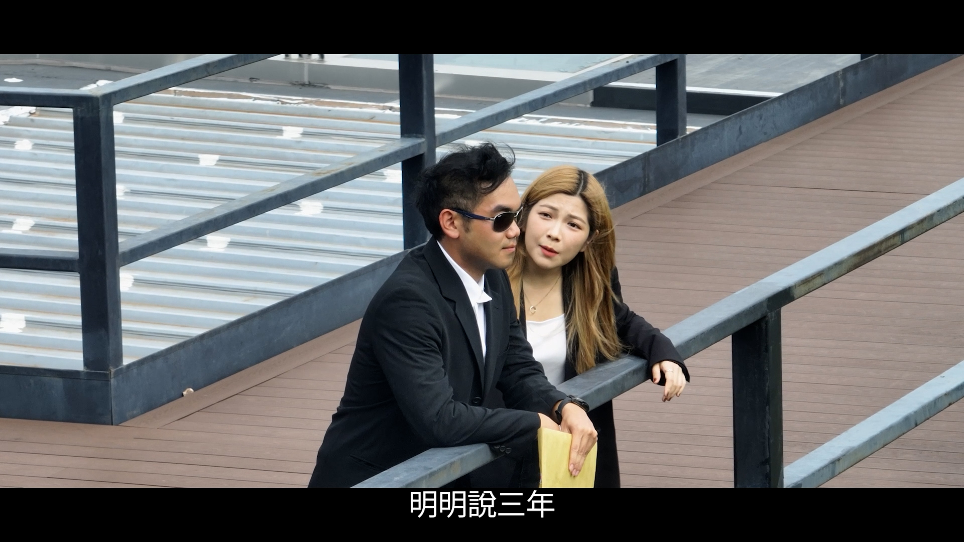 A man in a black suit and sunglasses leans on a railing, holding a paper, while a woman in a black jacket stands beside him, looking at him. Chinese subtitles are at the bottom of the image.